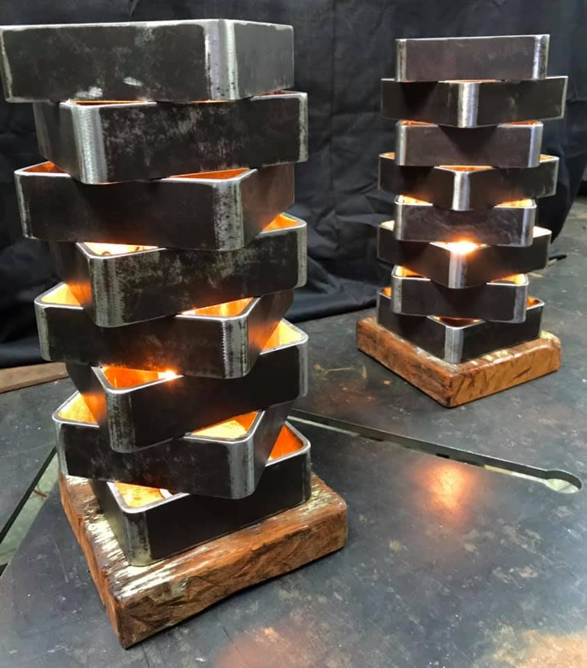 Two metal art sculptures shaped like vertically stacked, zigzagging pieces on wooden bases, with light glowing from inside. One sculpture is in the foreground, and another is in the background on a dark surface.