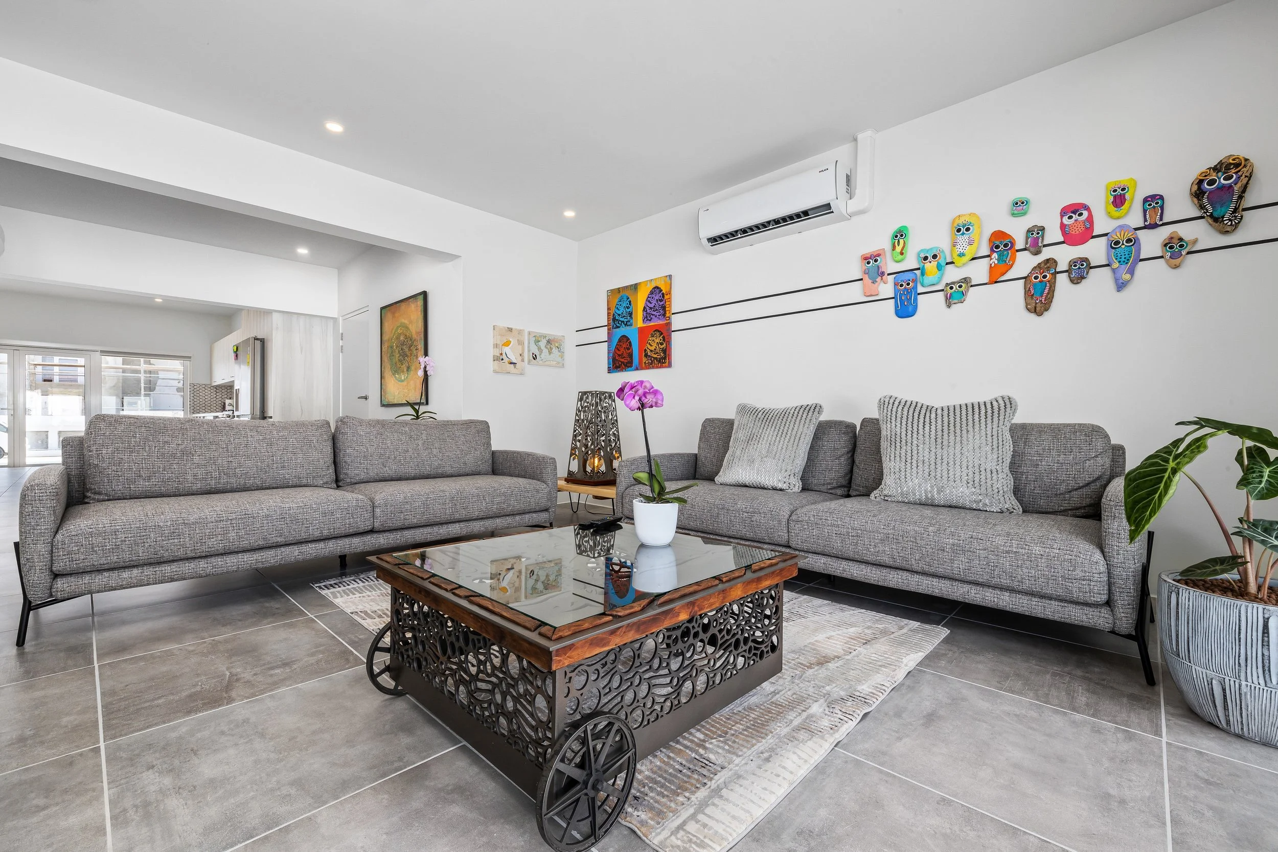 Modern living room with two gray sofas, a coffee table with a glass top and intricate black base, potted orchid, and colorful owl artwork on the wall.