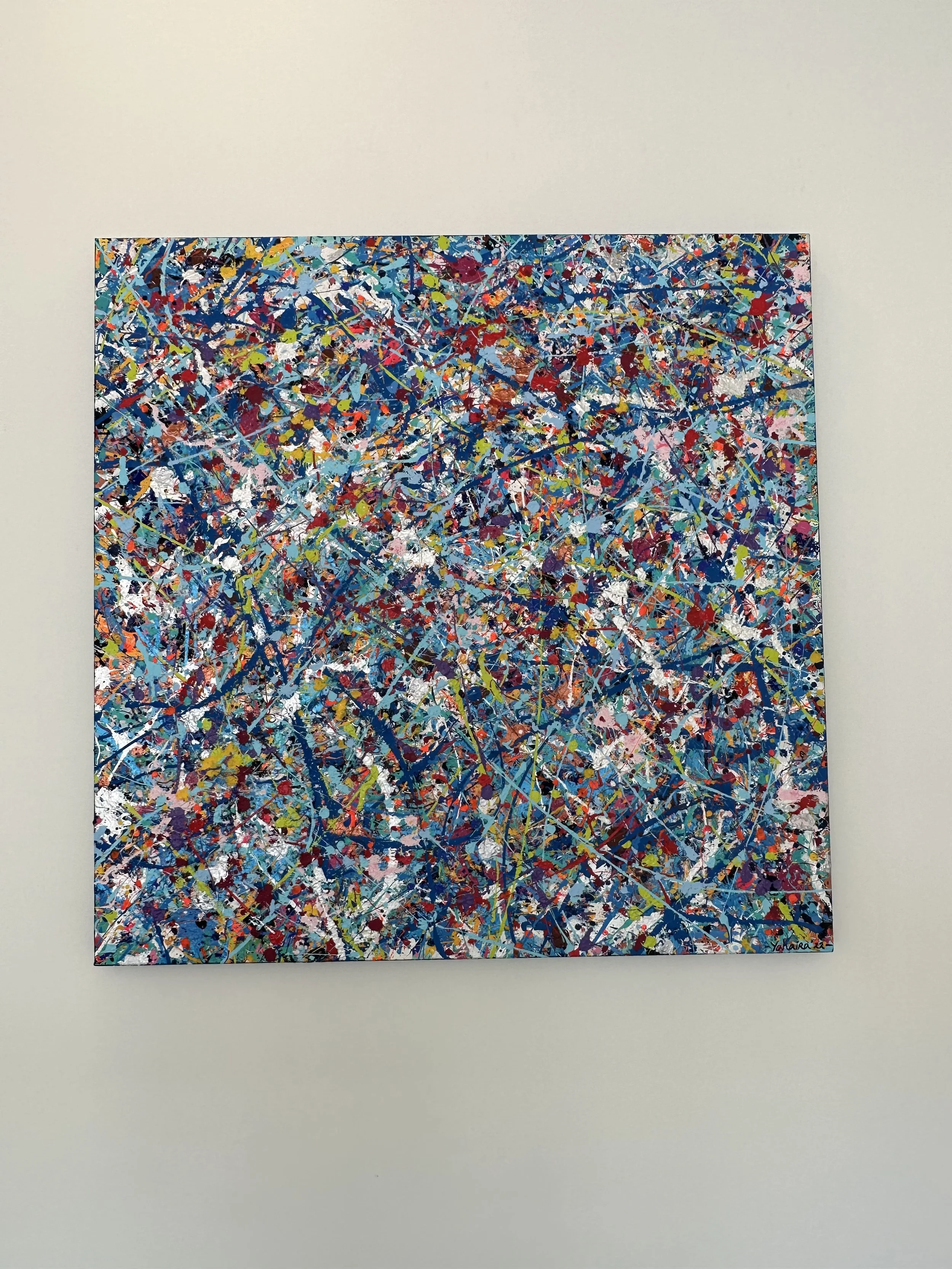 Abstract expressionist painting with multicolored splatters and lines on a square canvas, mounted on a plain white wall.