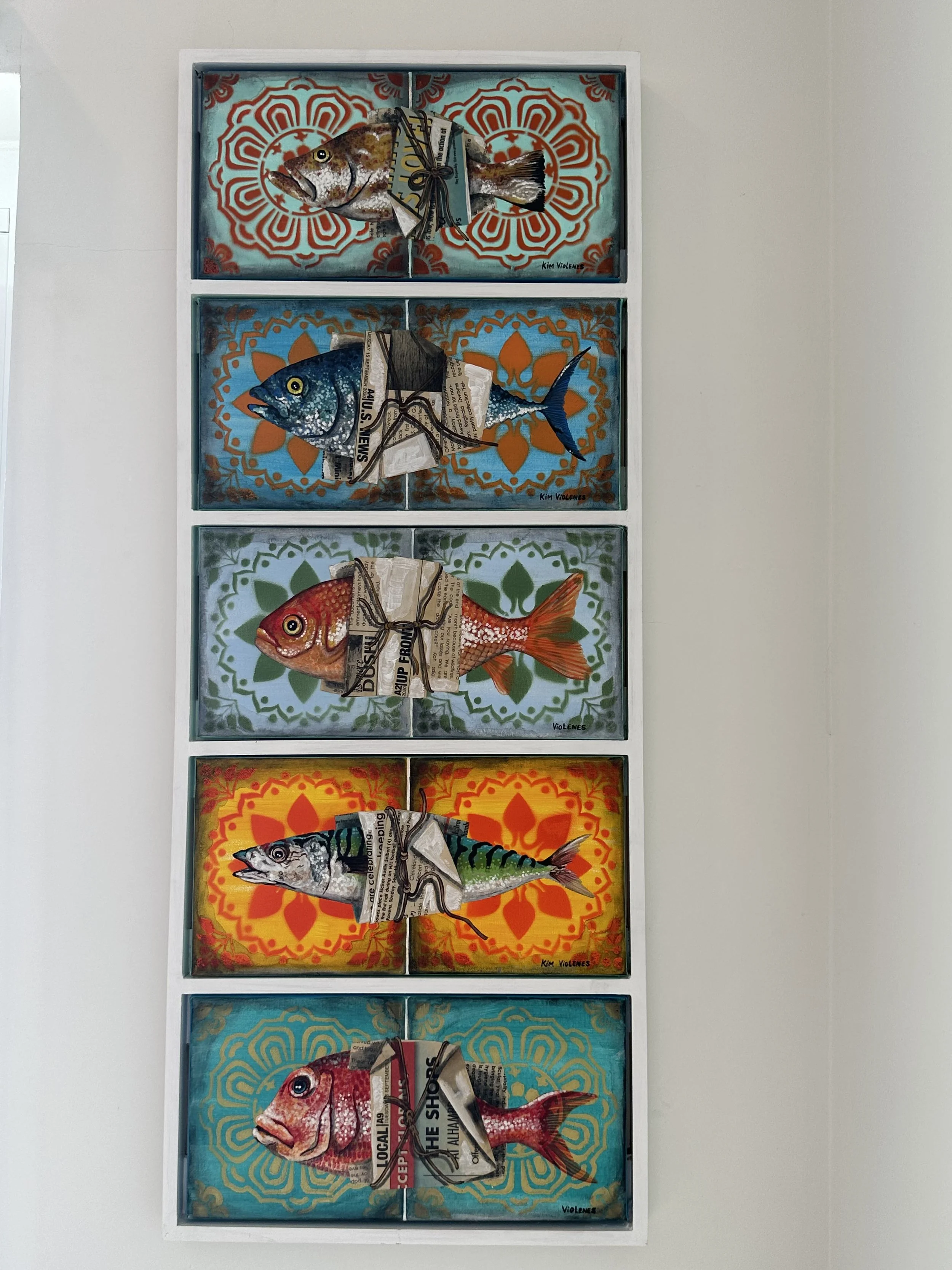 Five framed mixed media art pieces of fish, each with vibrant backgrounds and bundled with newspaper and string, arranged vertically on a wall.