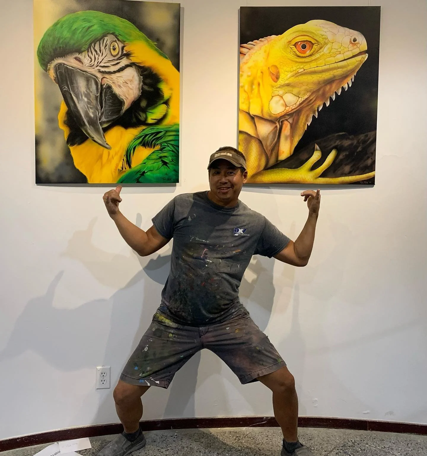 A man in paint-splattered clothes makes a flexing pose, standing in front of two animal paintings on a gallery wall, one of a parrot and one of a yellow lizard.