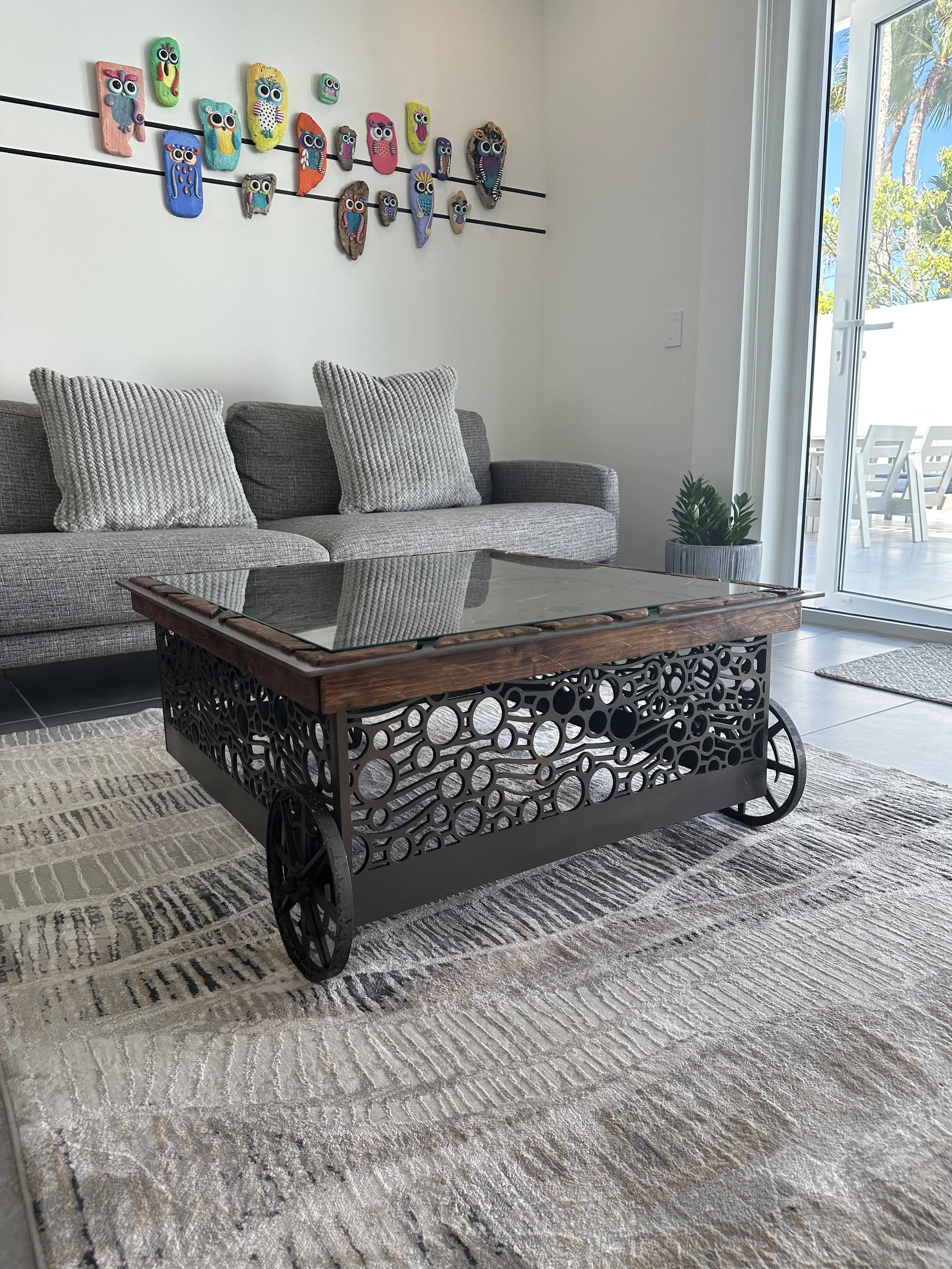 Living room with a gray sofa with textured pillows, a decorative cart coffee table with a glass top and circular cutout metal sides, a patterned area rug, artwork of colorful owl faces on the wall, potted plant near a sliding glass door leading outdo