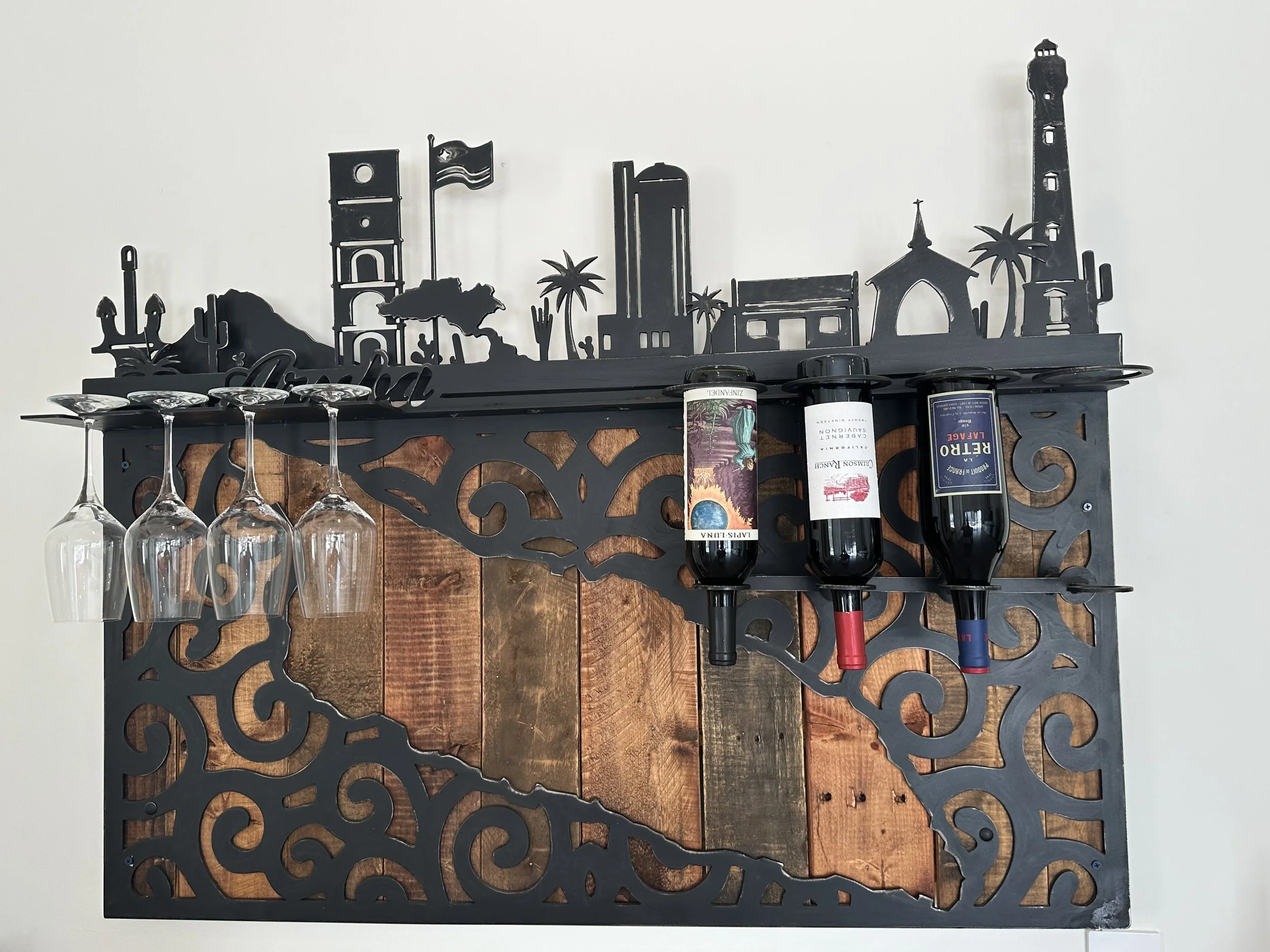 Wine rack with four upside-down wine glasses and three bottles of wine, decorative black metal landscape silhouette on top, and a white wall background.