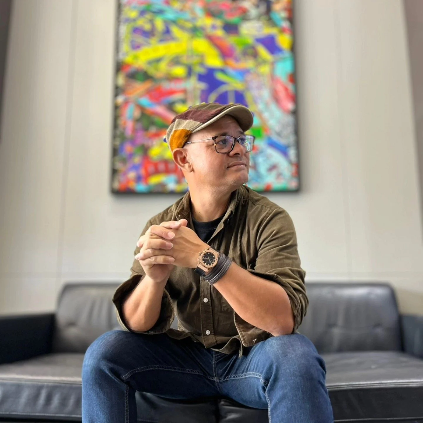 A man sitting on a black leather sofa in an art gallery with a colorful abstract painting hanging behind him. He is wearing glasses, a brown cap, a brown shirt with rolled-up sleeves, blue jeans, and a watch. His hands are clasped together as he looks to the side.