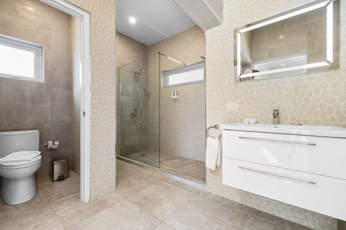 Modern bathroom with a toilet, a walk-in shower with glass door, a wall-mounted mirror with lights, and a white vanity with a sink.