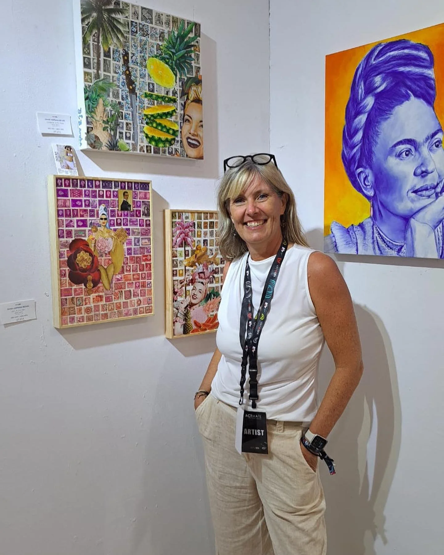 A woman smiling at an art gallery, standing in front of colorful artwork including portraits and collage-style pieces on a white wall.