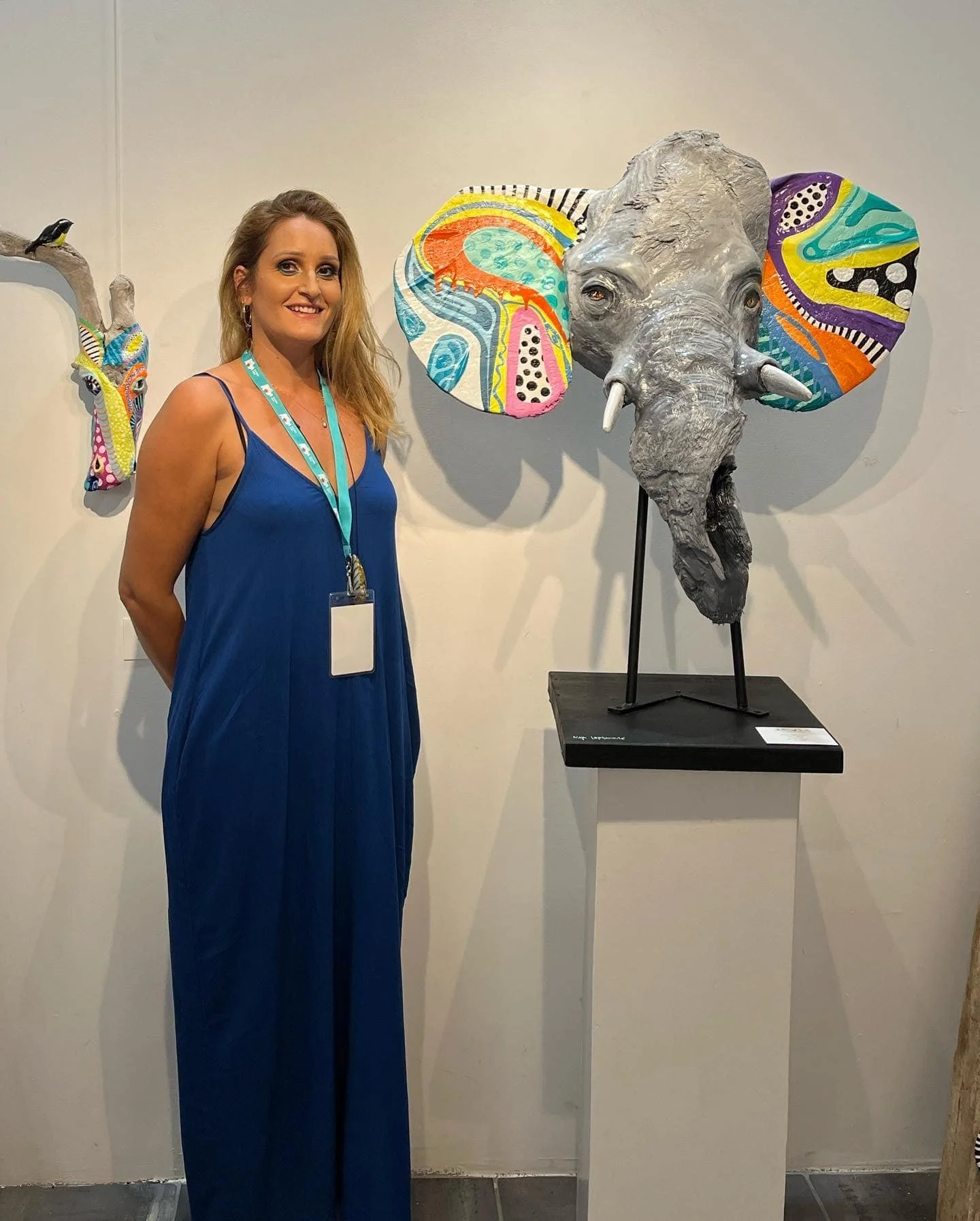 A woman in a blue jumpsuit standing next to a sculpture of an elephant's head with colorful, abstract painted ears, in an art gallery.