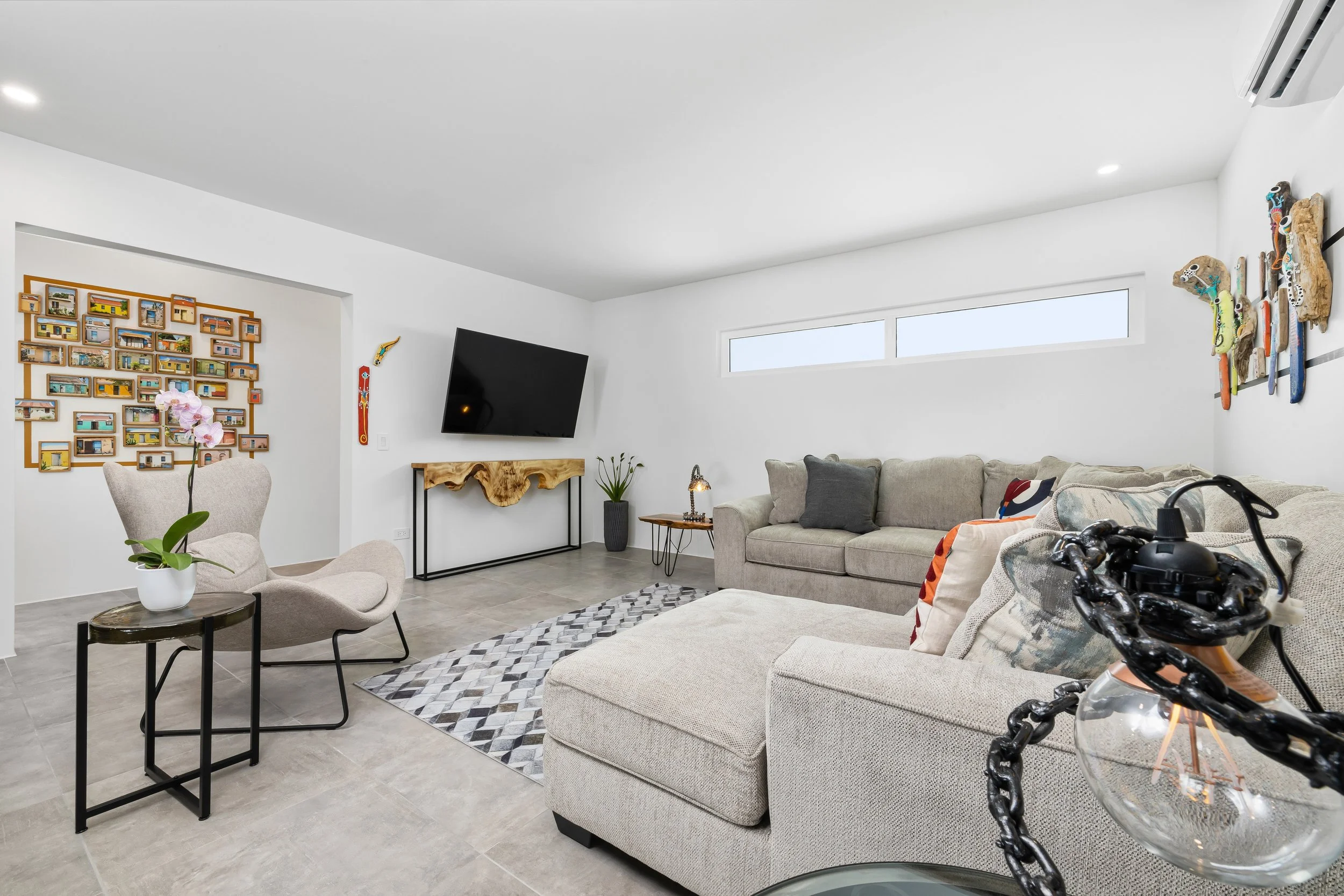 Modern living room with beige sofas, a patterned rug, wall art, a mounted TV, a small table with a lamp, a chair with a flowering plant, and decorative objects.