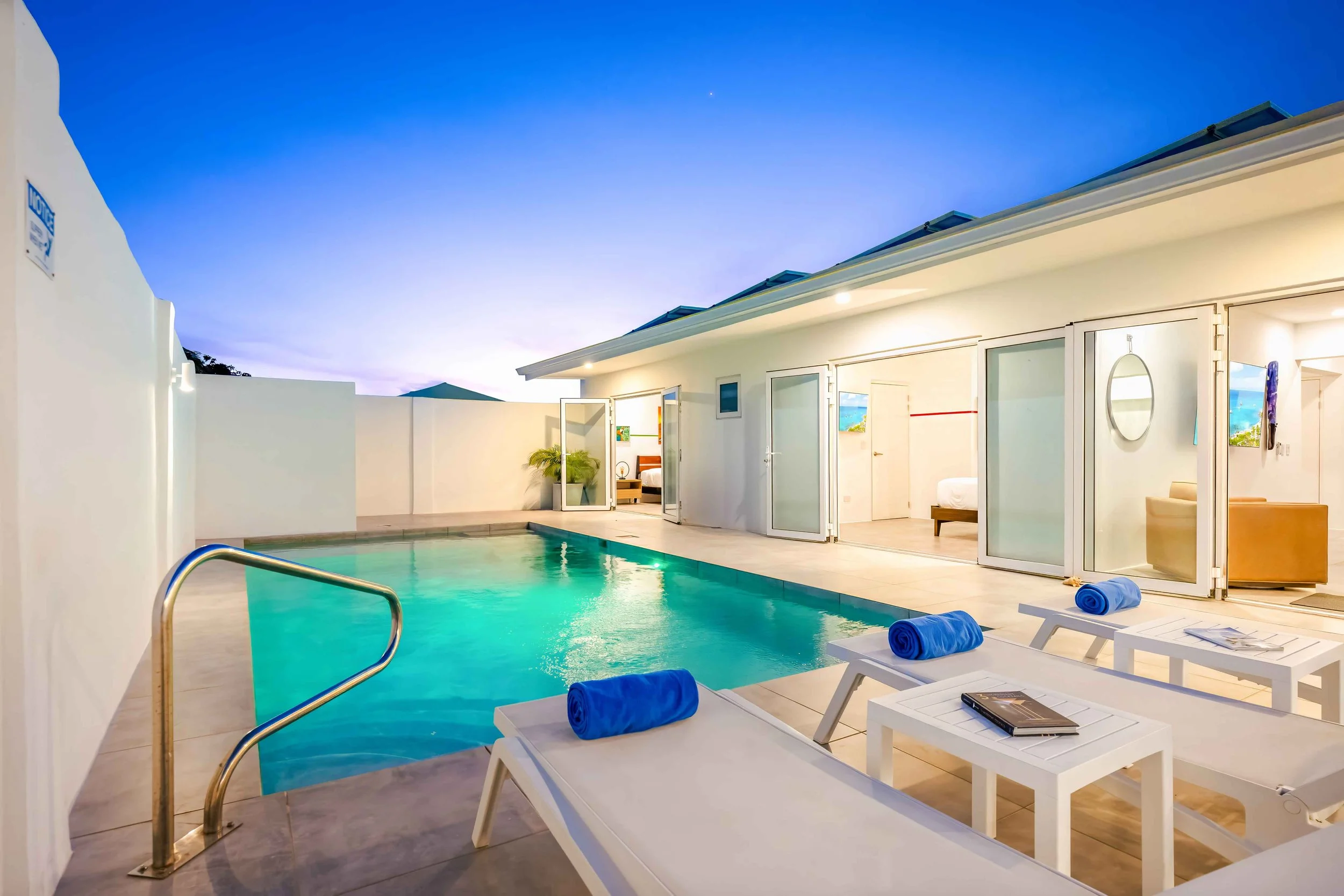 A backyard pool area with three lounge chairs and rolled towels, surrounded by white walls and a modern house with glass doors, under a blue evening sky.