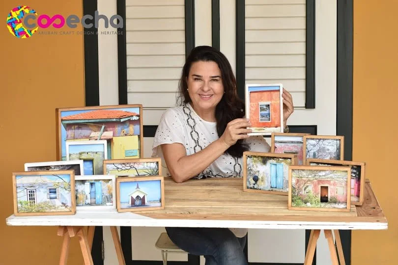 A woman sitting at a wooden table displaying framed photographs of colorful buildings and landscapes. She is smiling and holding up one of the frames. The background features a window with closed blinds and a two-tone wall.