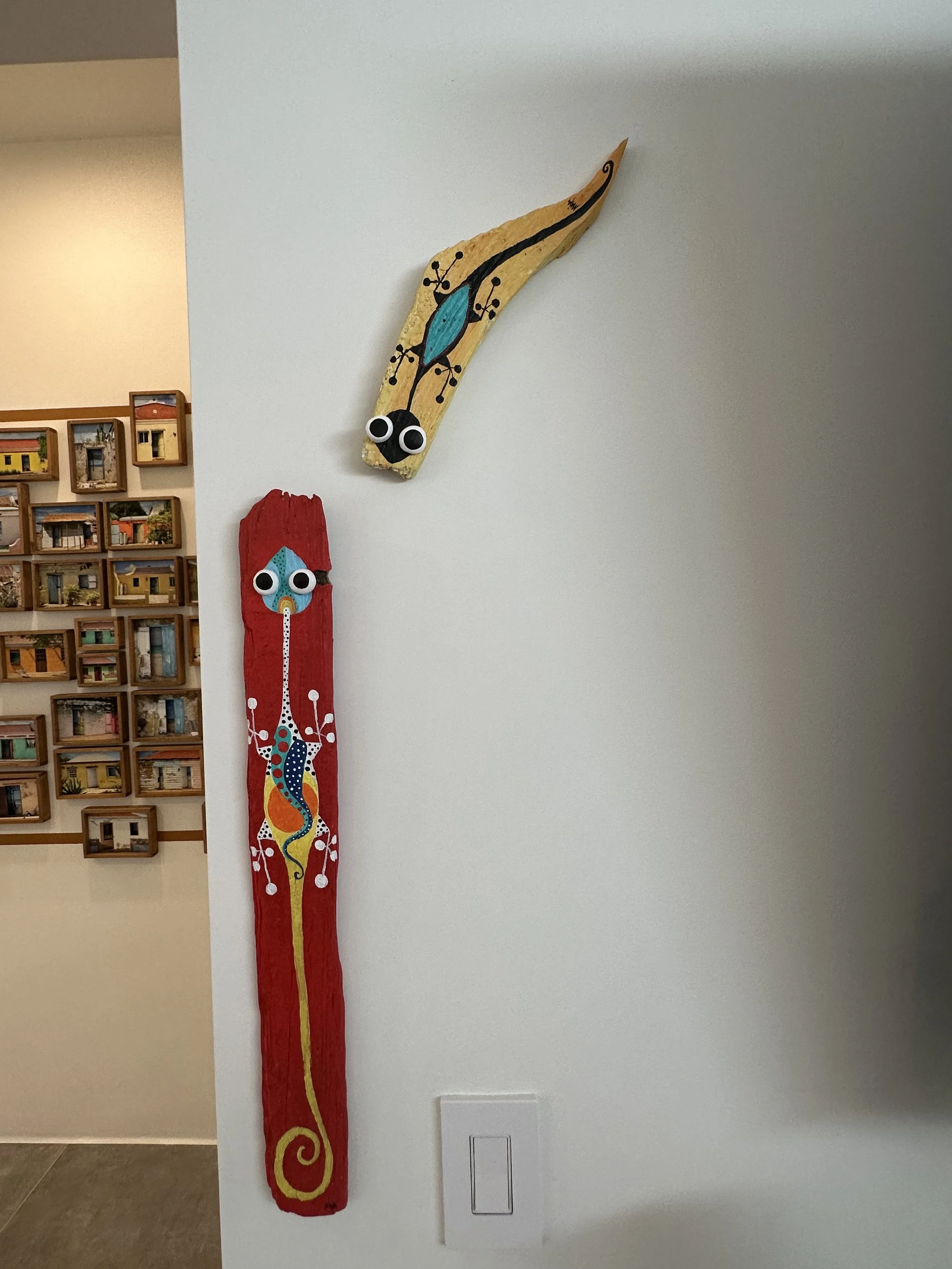 Colorful wooden wall art with googly eyes, one shaped like a yellow and black lizard or gecko, and the other like a red and blue abstract creature, displayed on a white wall.