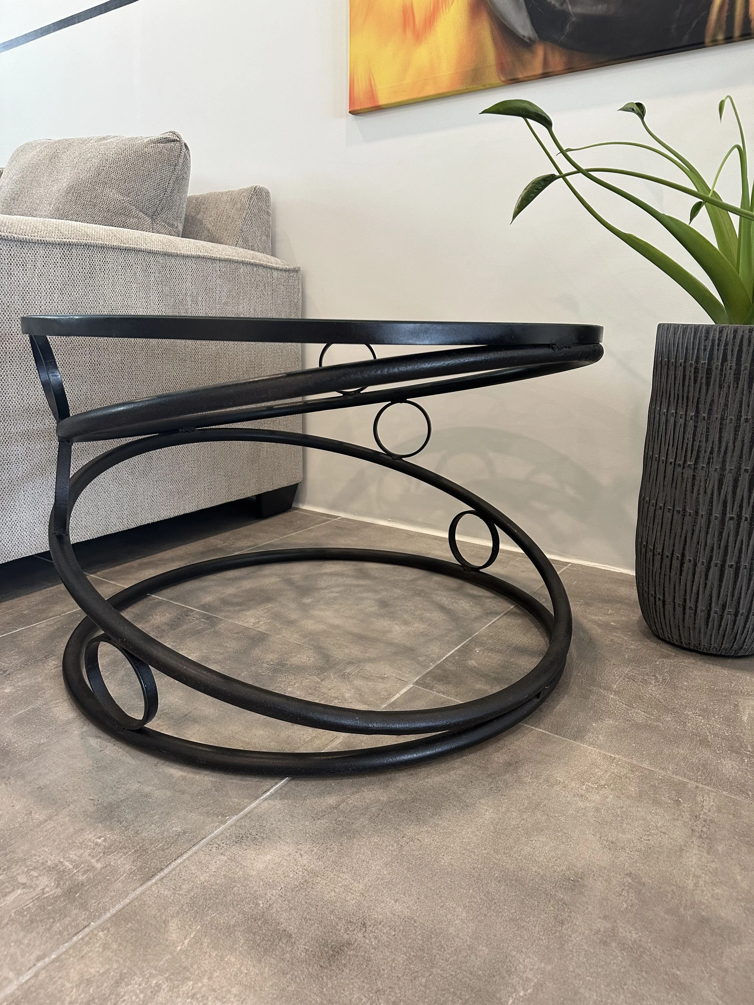 Black metal side table with circular leg design on a grey tiled floor, beside a grey couch and a tall black textured planter with a green plant, in a modern living room.