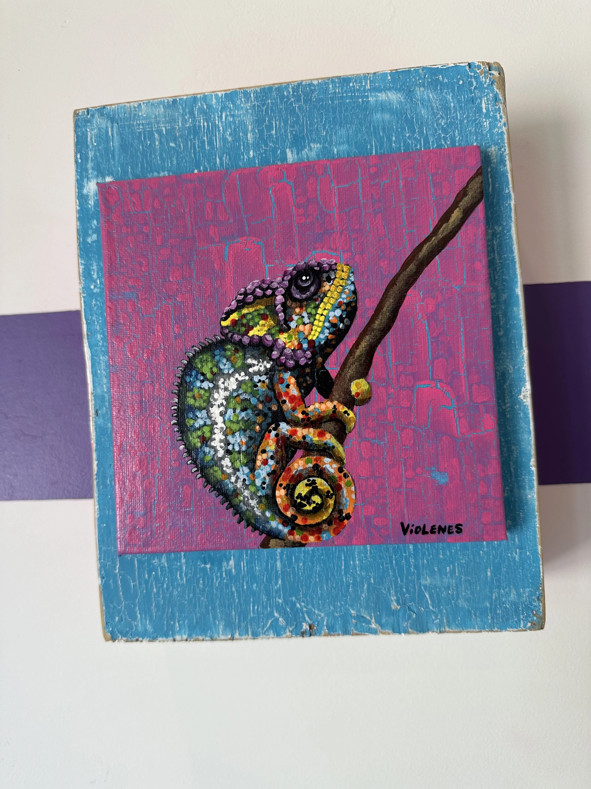 Colorful painted chameleon perched on a branch with a pink background and a blue border, signed by Violenes.
