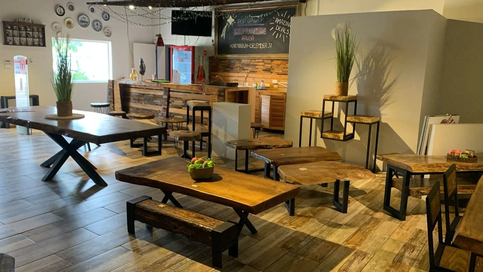Interior of a rustic restaurant or cafe with wooden tables and chairs, potted plants, wall decoration, and a chalkboard menu.