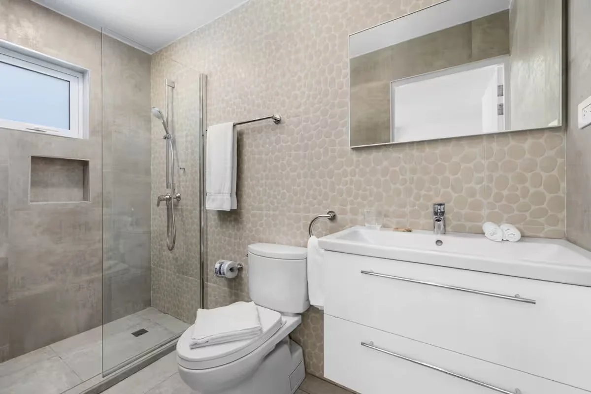 Modern bathroom with a walk-in shower, toilet, and white vanity sink with a large mirror above. The walls and floor are tiled in neutral tones, with small pebble-like tiles on the wall behind the sink. There is a small window near the shower.