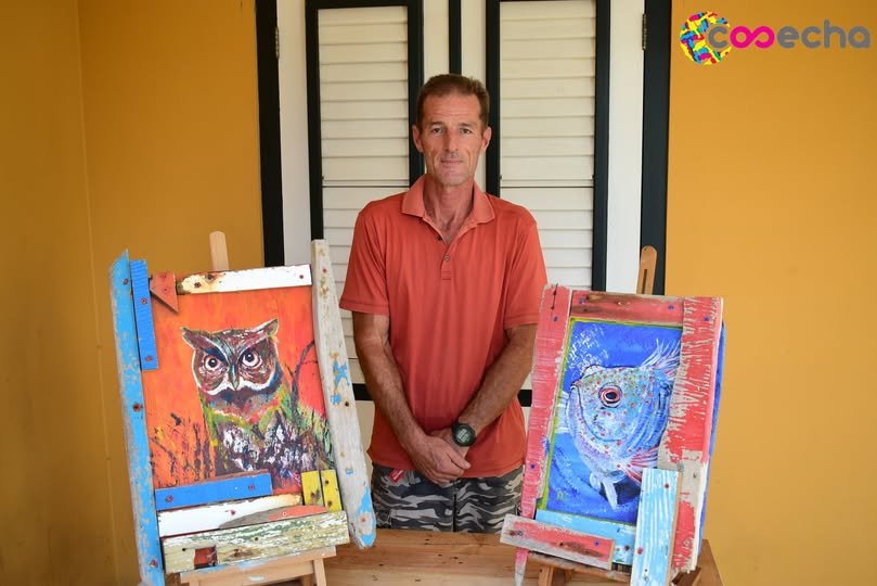 A man wearing a red shirt standing behind a table with two colorful paintings of animals on easels, set against a yellow wall with closed window shutters.