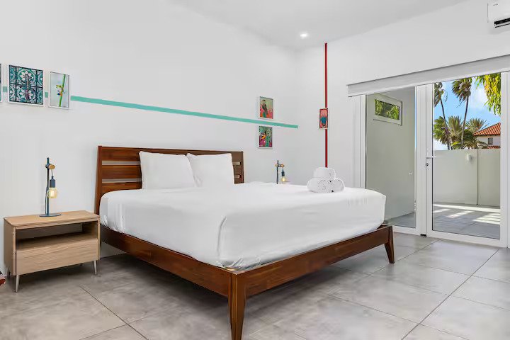 Modern bedroom with a wooden bed, white bedding, and two nightstands with lamps. Artwork decorates the white walls, and a sliding glass door leads to a balcony with palm trees visible outside.