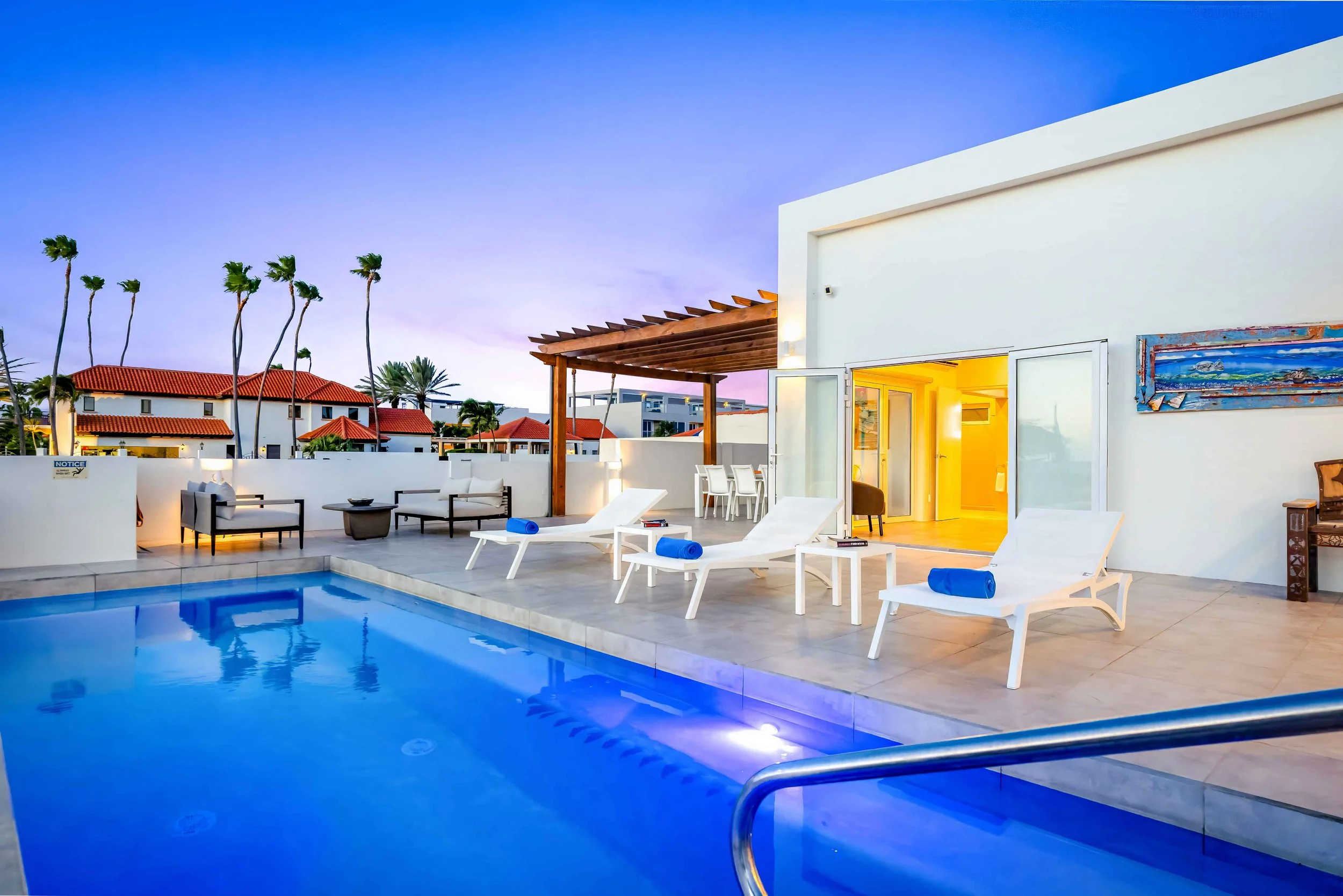 Swimming pool area with white lounge chairs and blue towels, outdoor seating, and a patio with a wooden pergola under a purple dusk sky.