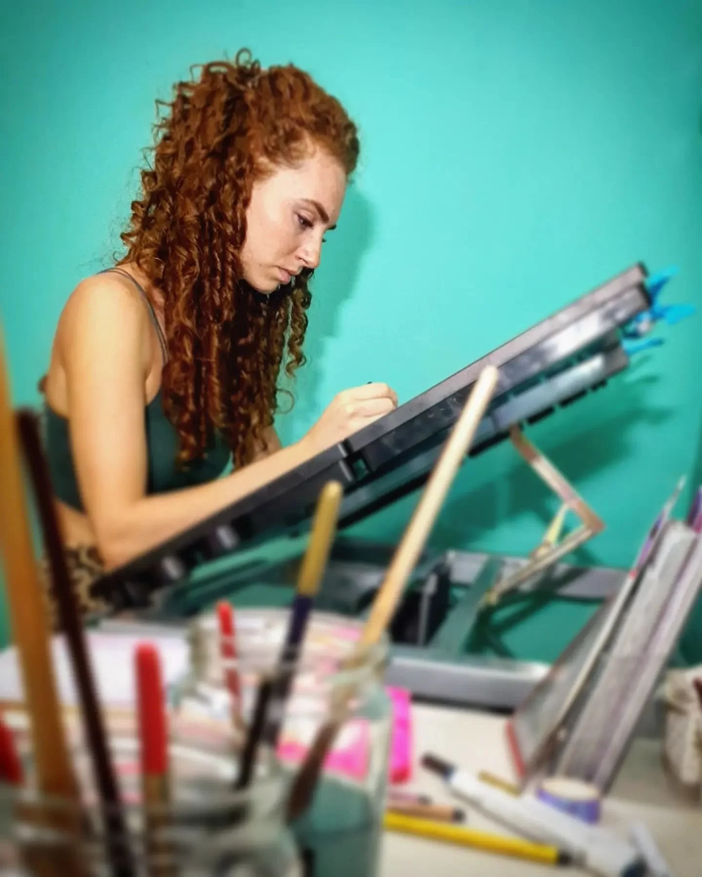 A woman with long curly red hair working on a digital drawing tablet surrounded by art supplies on a desk.