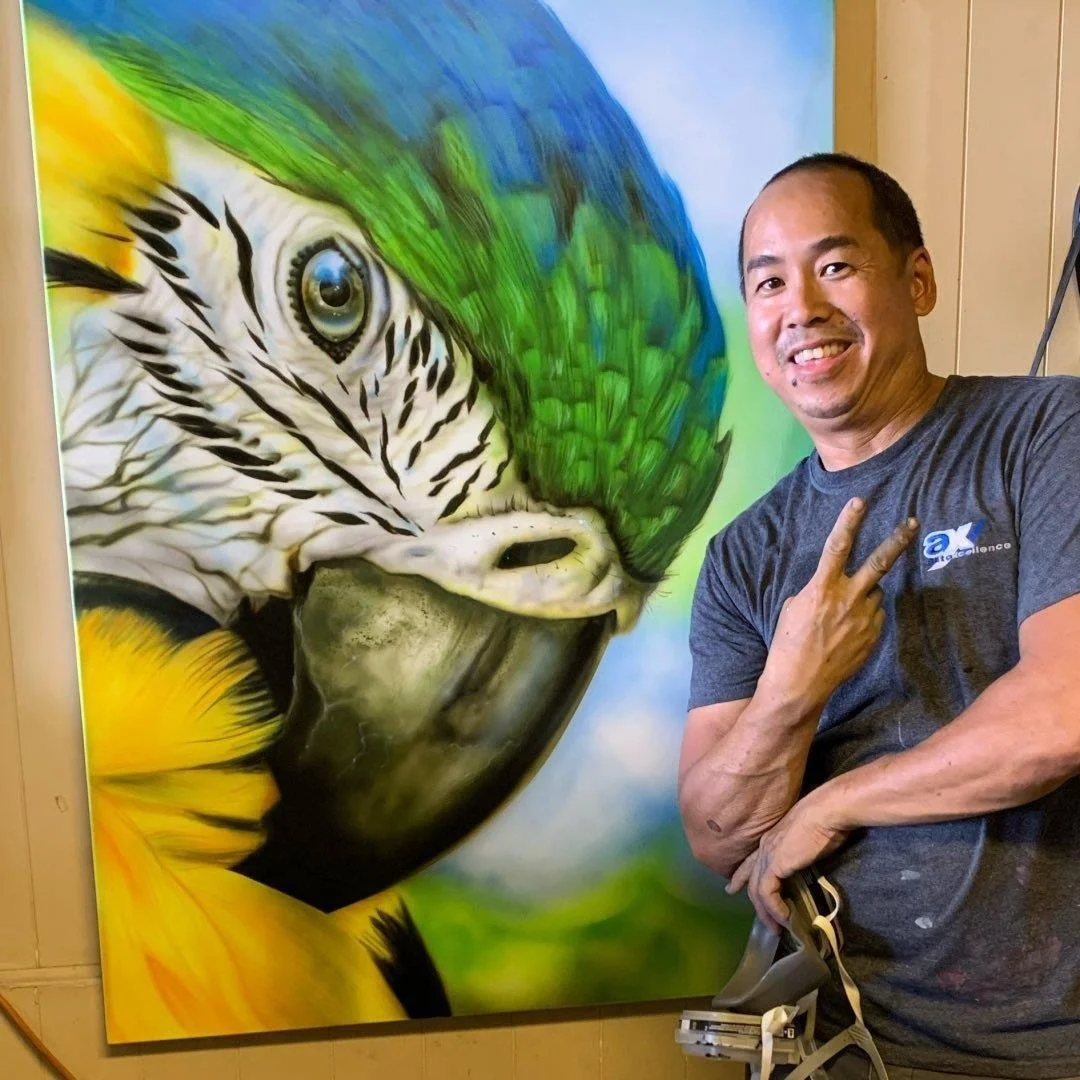 A man smiling at a colorful painting of a parrot, with the man making a peace sign.
