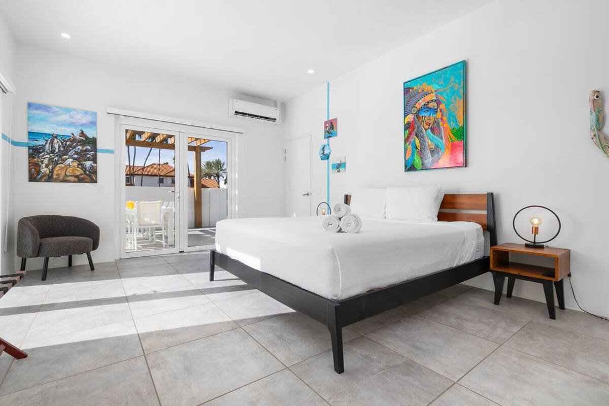 Modern bedroom with a large bed, white bedding, colorful artwork on white walls, a nightstand with a lamp, and a sliding glass door leading to a balcony with outdoor furniture.