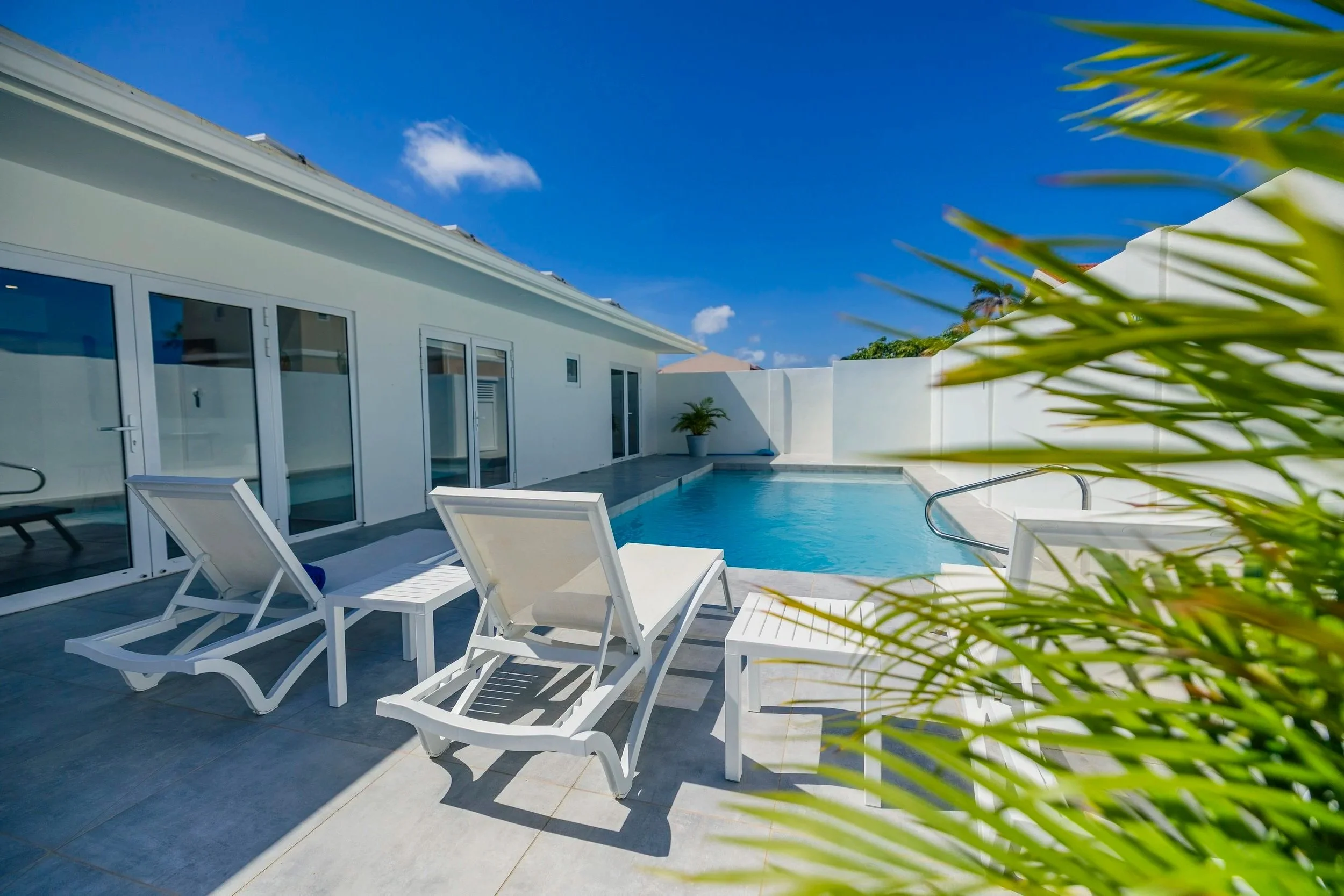 A private outdoor pool area with white lounge chairs on a tiled deck, a white modern house with large glass windows and doors, sunny weather with clear blue sky, and some greenery in the foreground.