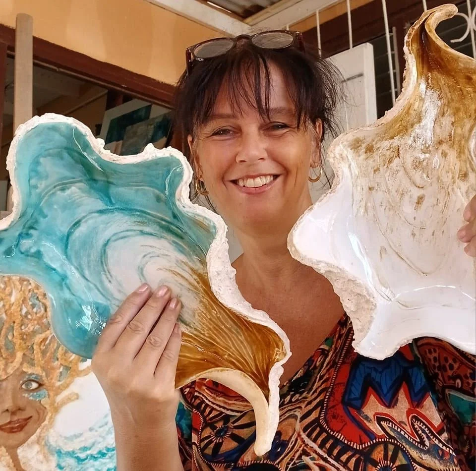 Woman smiling while holding two large, colorful seashell-shaped objects, one with swirling blue and white patterns and the other with brown and white patterns.