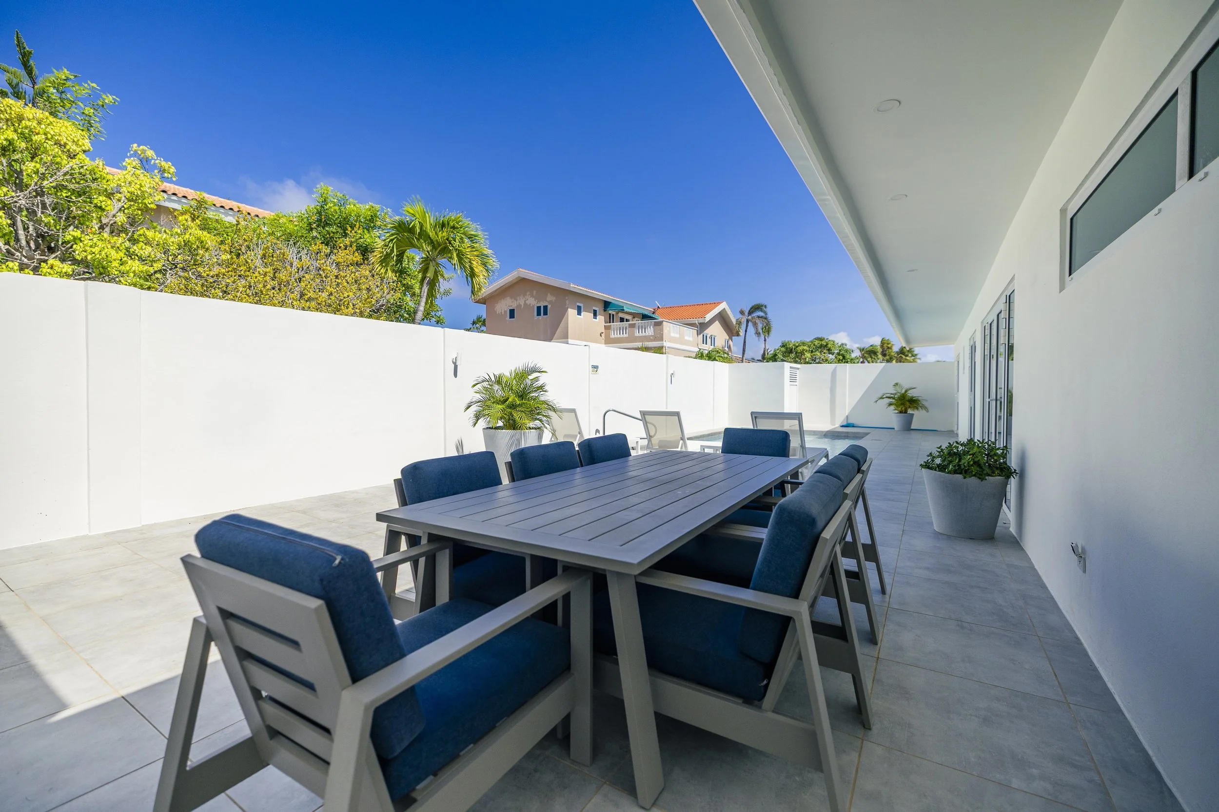 Modern outdoor patio with a large dining table and blue cushioned chairs, potted plants, white wall, and neighboring houses visible against a clear blue sky.