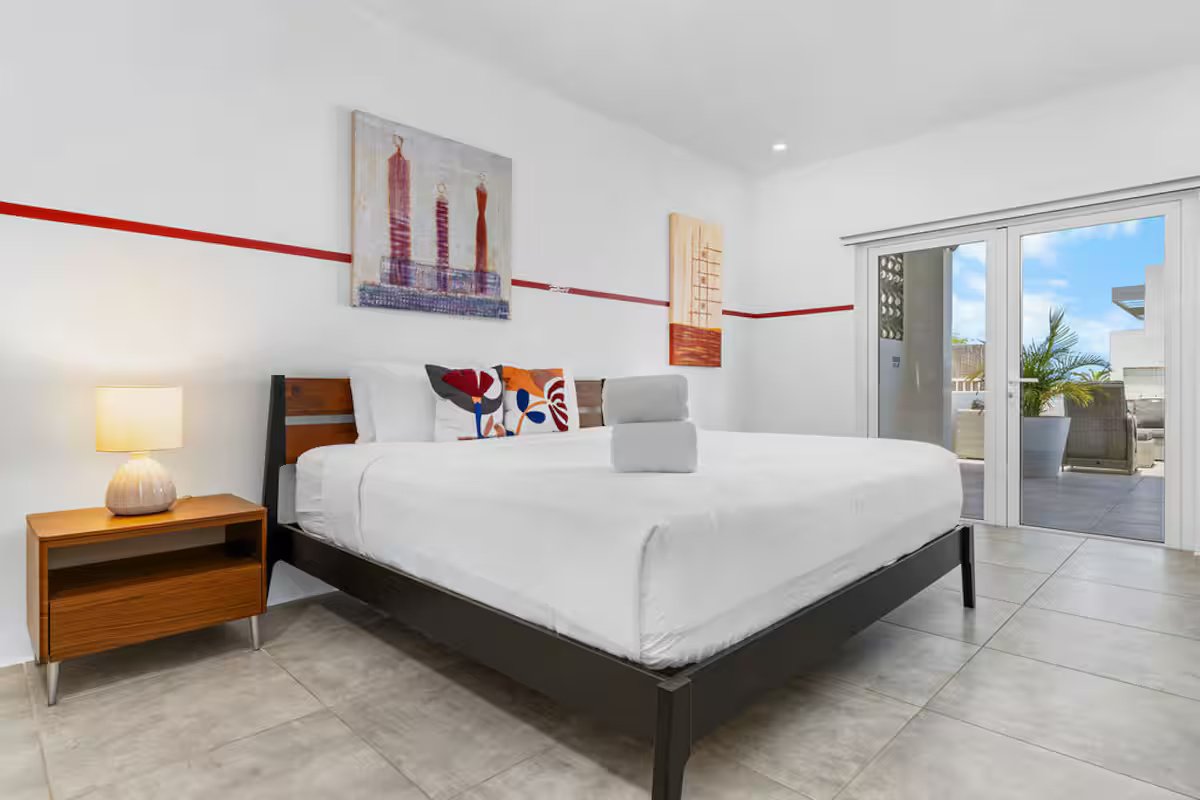 Modern bedroom with a large bed, colorful pillows, a wooden nightstand with a lamp, artwork on the wall, and sliding glass door leading to a balcony with outdoor furniture and palm trees.