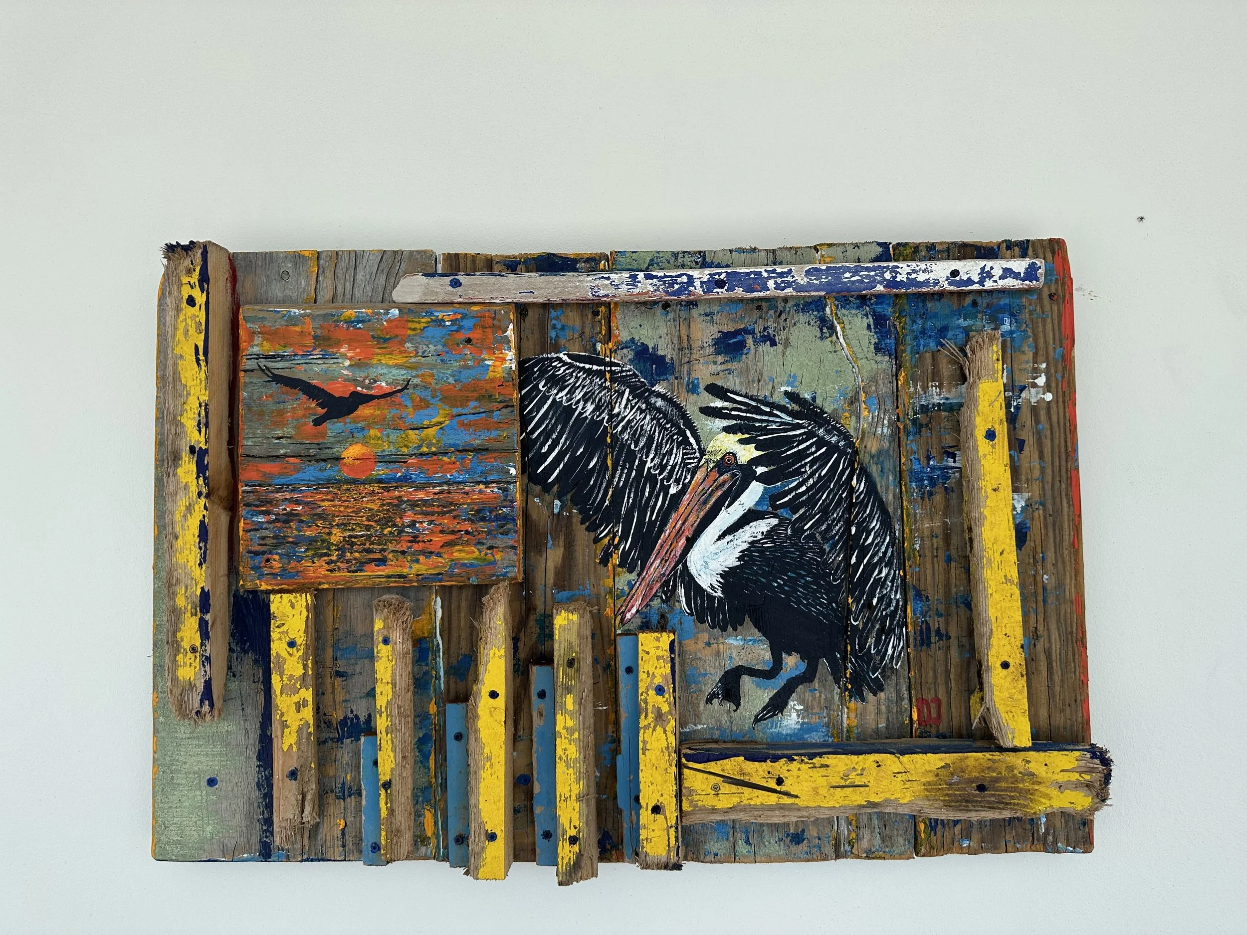 Mixed media artwork on wooden pallets featuring a painted sunset with a bird and a painted black and white pelican.