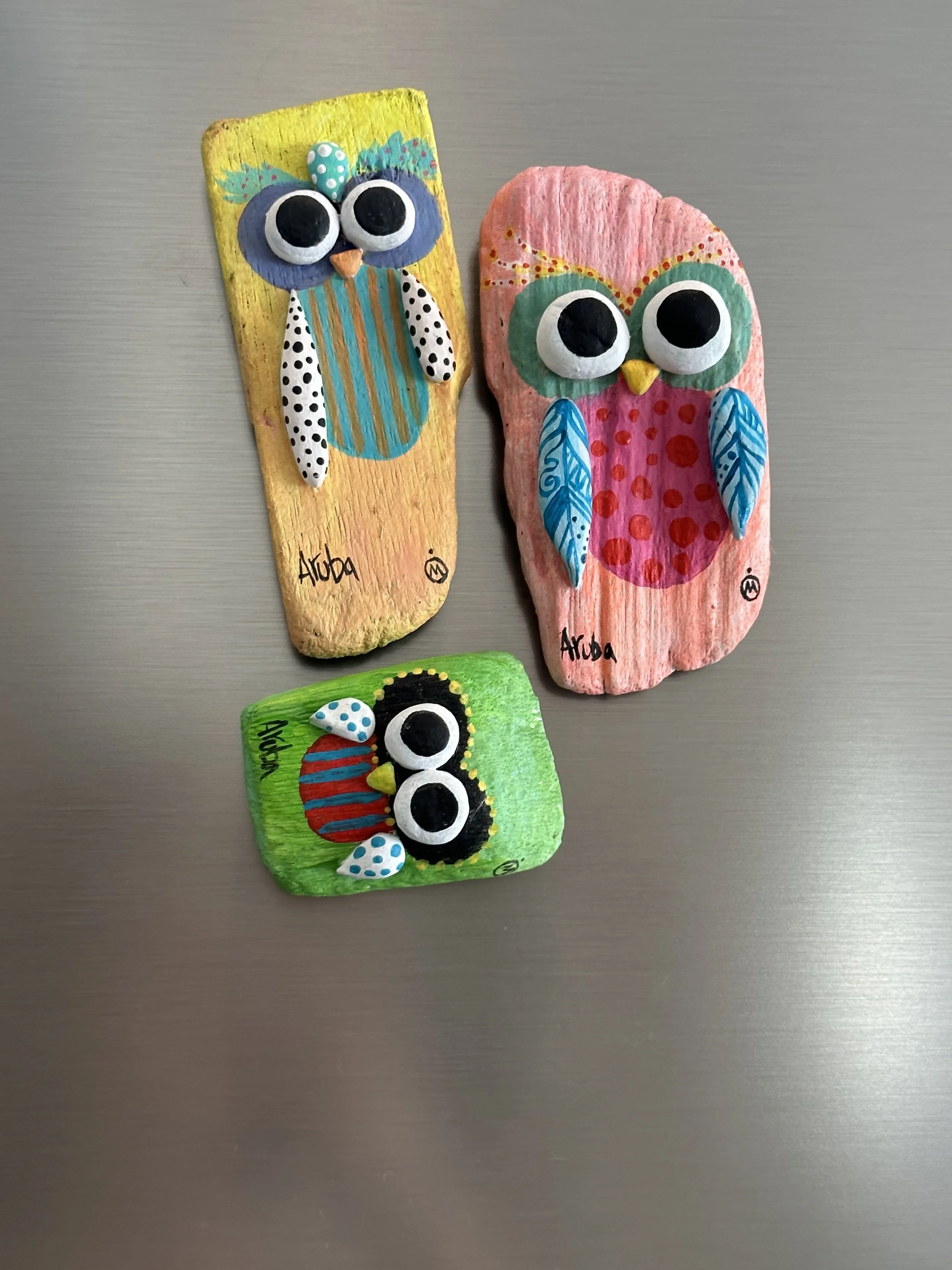 Three painted rocks decorated as colorful owl characters with large eyes, beak, and wings, placed on a metallic surface.