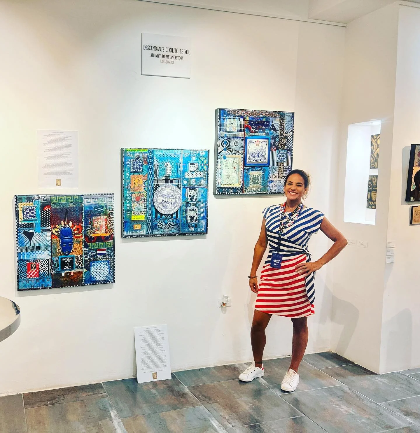 A woman standing in front of colorful, abstract artwork in a gallery, wearing a striped dress with an American flag pattern on the skirt, white sneakers, and smiling.