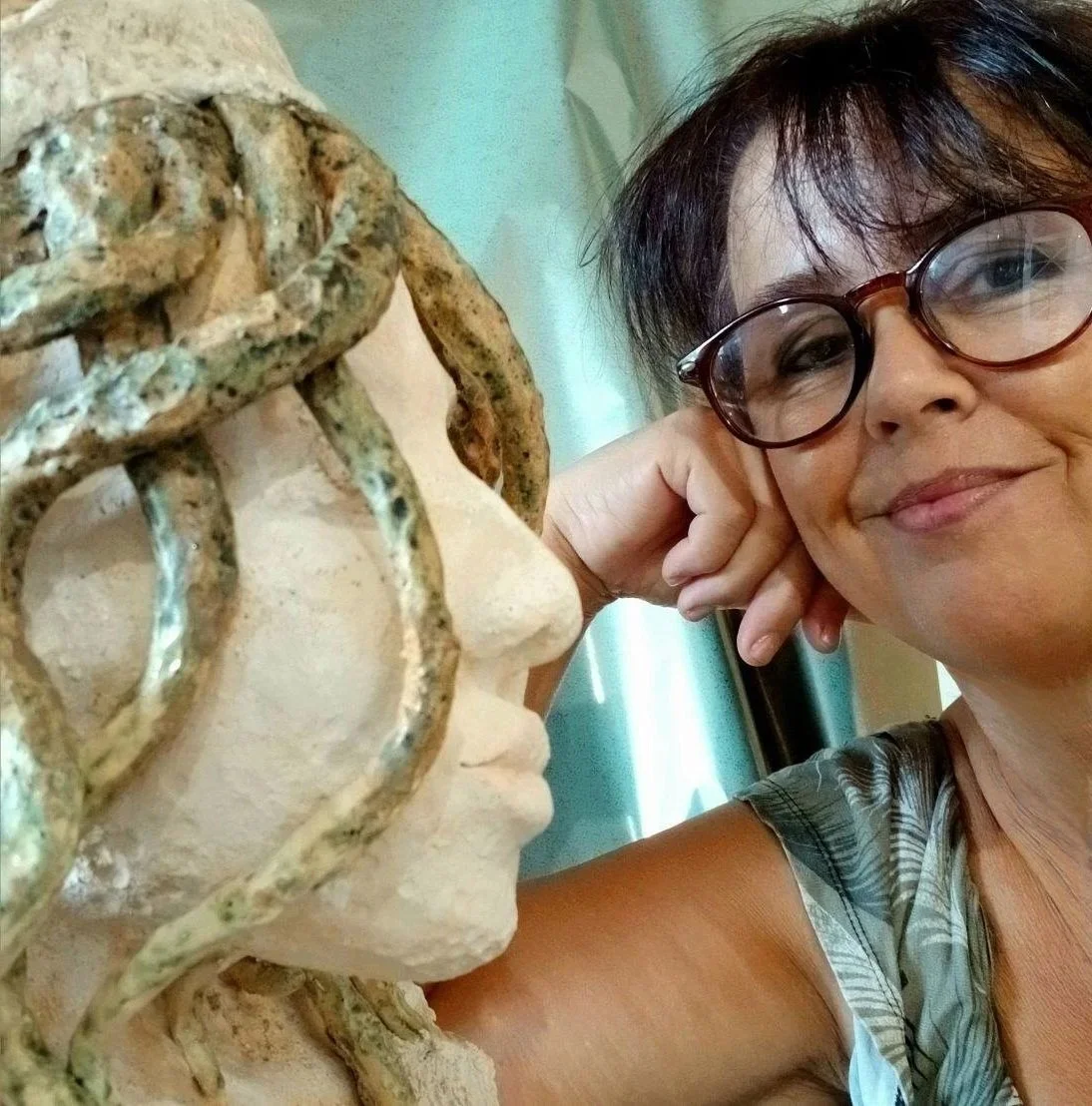 A woman with short dark hair and round glasses smiling, taking a selfie next to a stone sculpture of a person's face with wire hair.