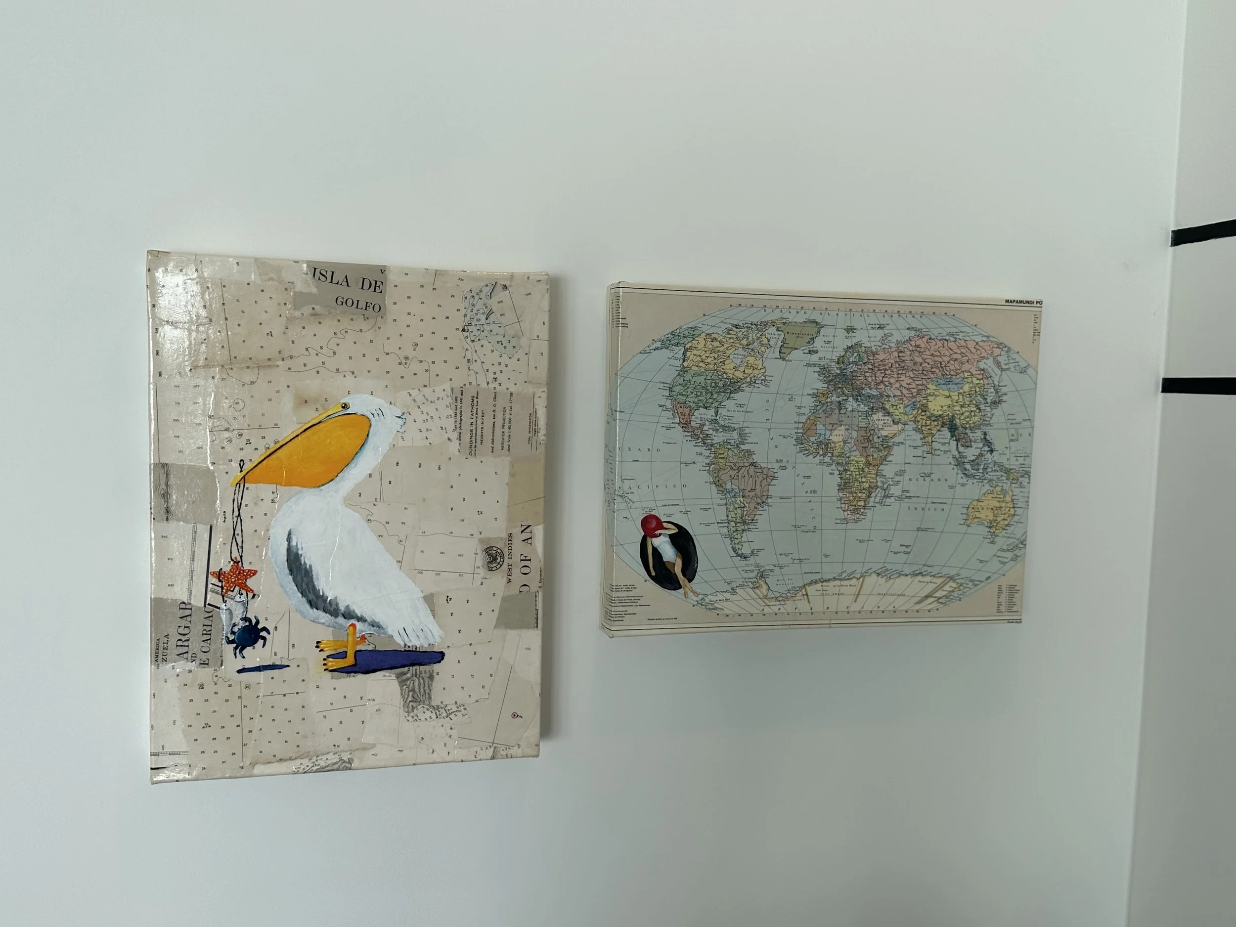 Two maps on a white wall. The left map is a collage with a painted pelican holding a starfish and a crab in front of a map background. The right map is a detailed world map with various countries and a piece of artwork of a bird on it.