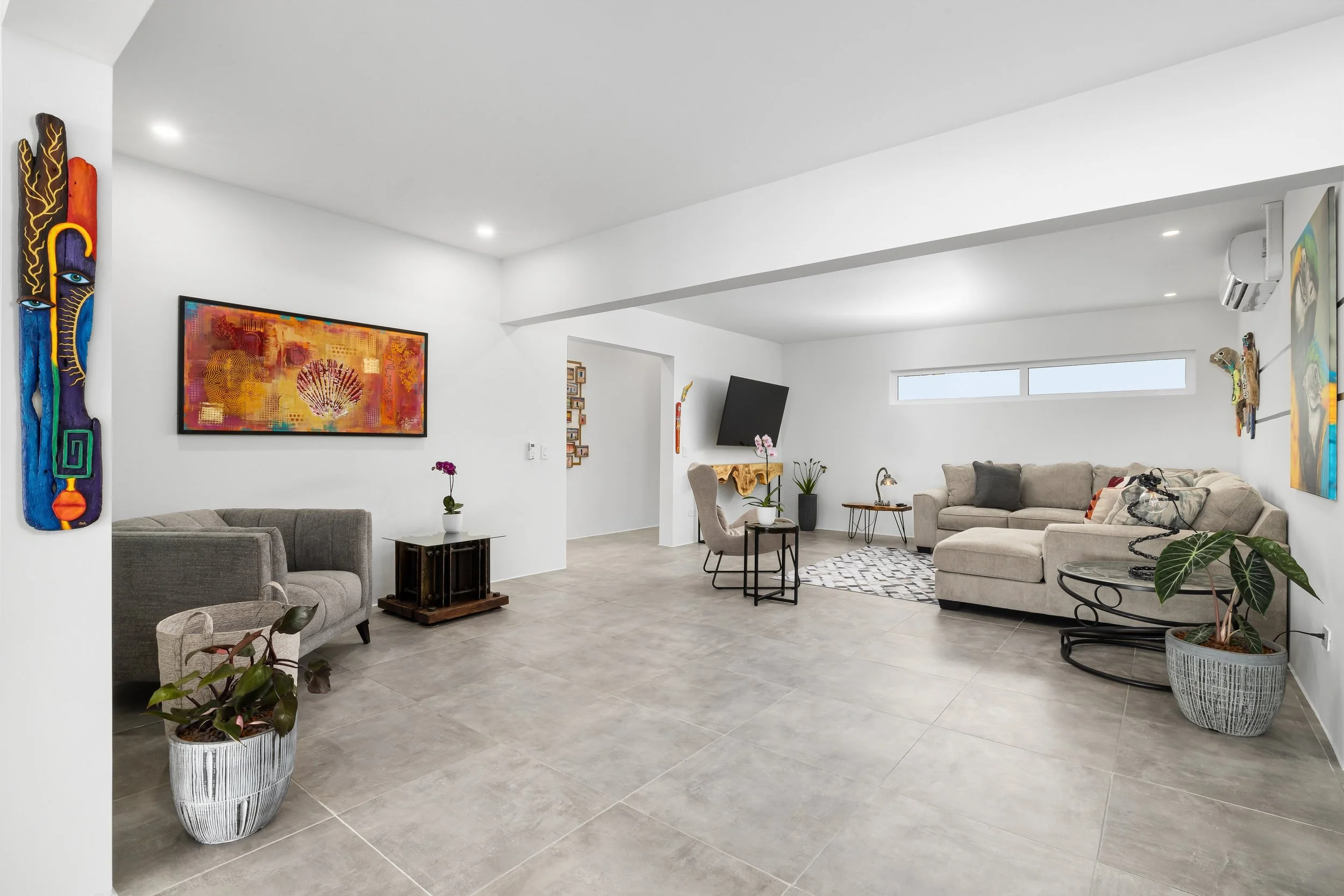 Modern living room with white walls, beige sectional sofa, black and white geometric rug, colorful abstract art, indoor plants, and wall-mounted TV.