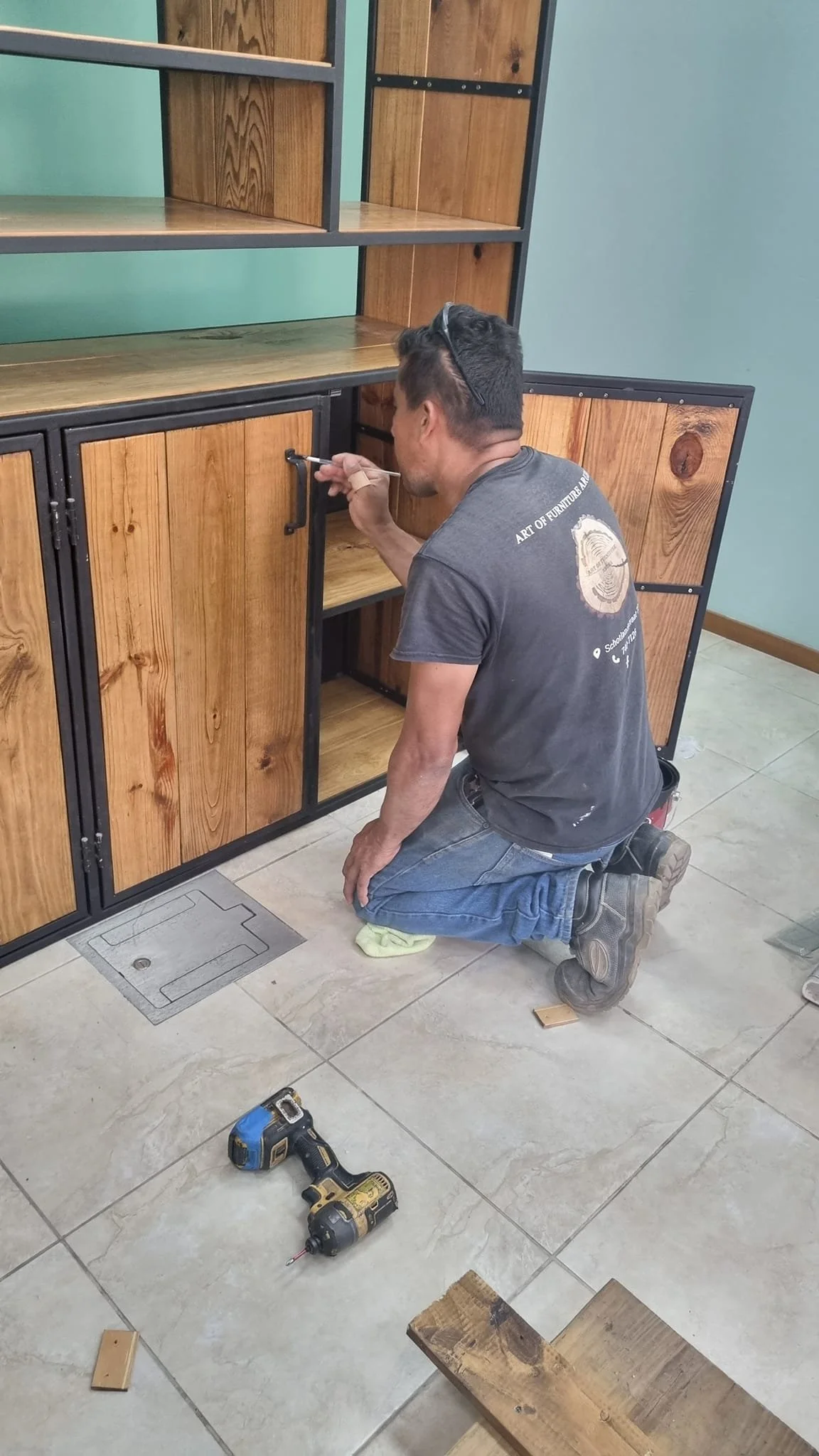 Man kneeling on the floor installing or repairing a wooden cabinet, with tools and hardware around him.