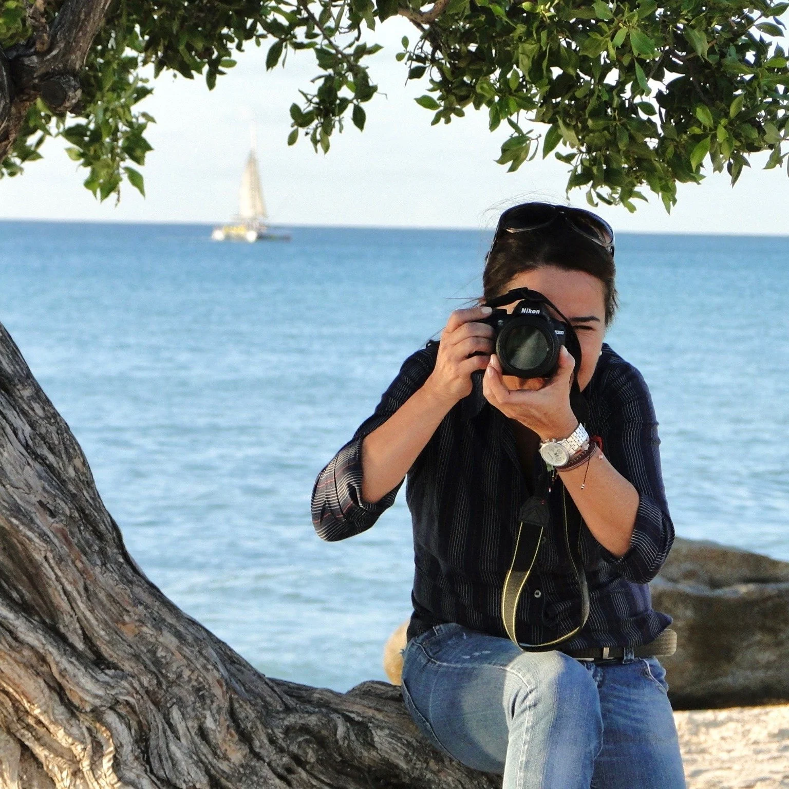 A woman sitting on a tree trunk by the water, taking a photo with a Nikon camera during daytime, with a sailboat in the distance on the water.
