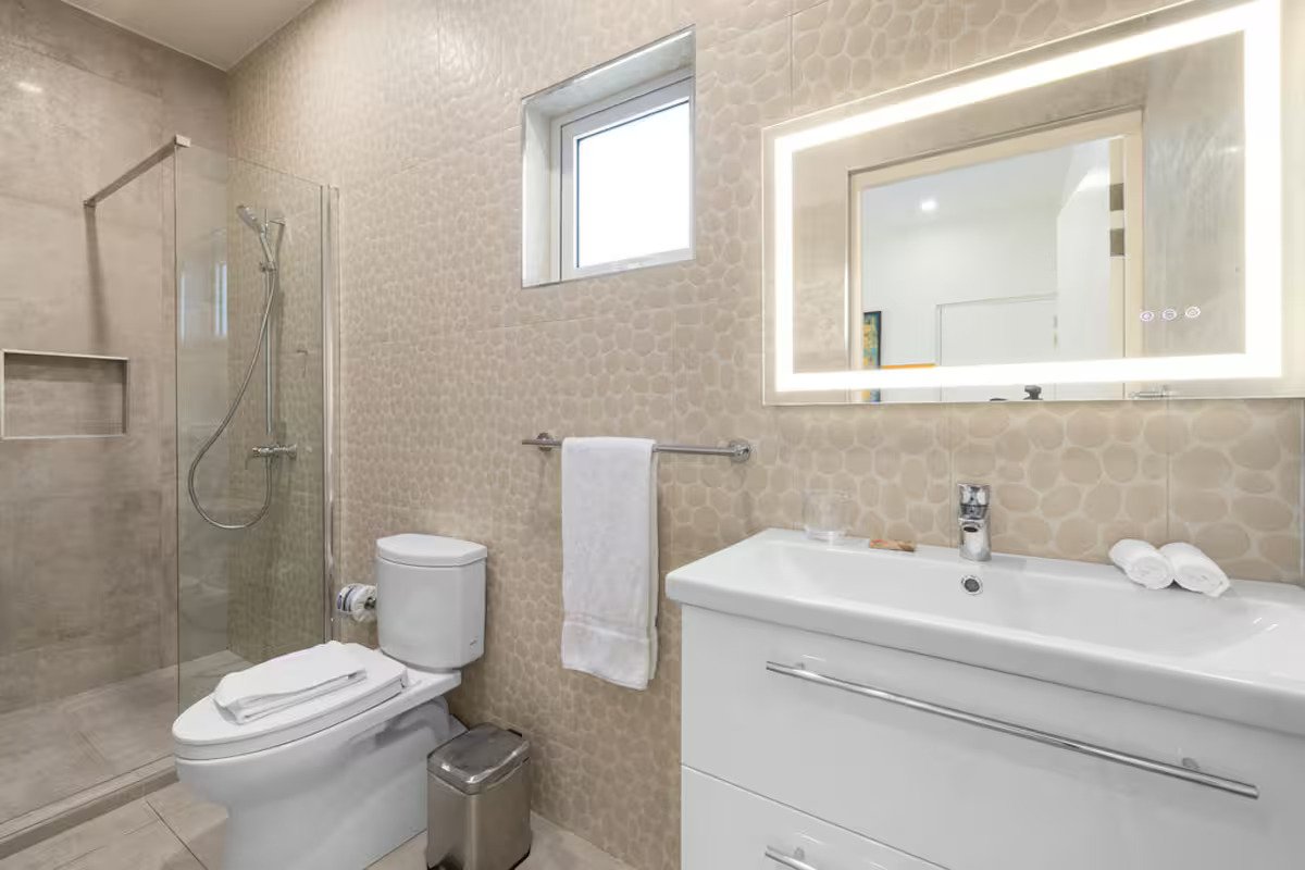 Modern bathroom with walk-in shower, white toilet, towel hanging on a rail, white vanity with a large illuminated mirror, small window, and beige textured tile walls.