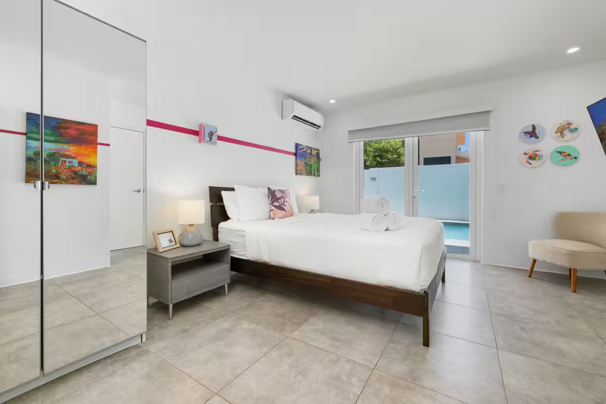 Modern bedroom with white walls, large bed with white linens, colorful artwork on the walls, a sliding glass door leading to a balcony, and light-colored tile flooring.