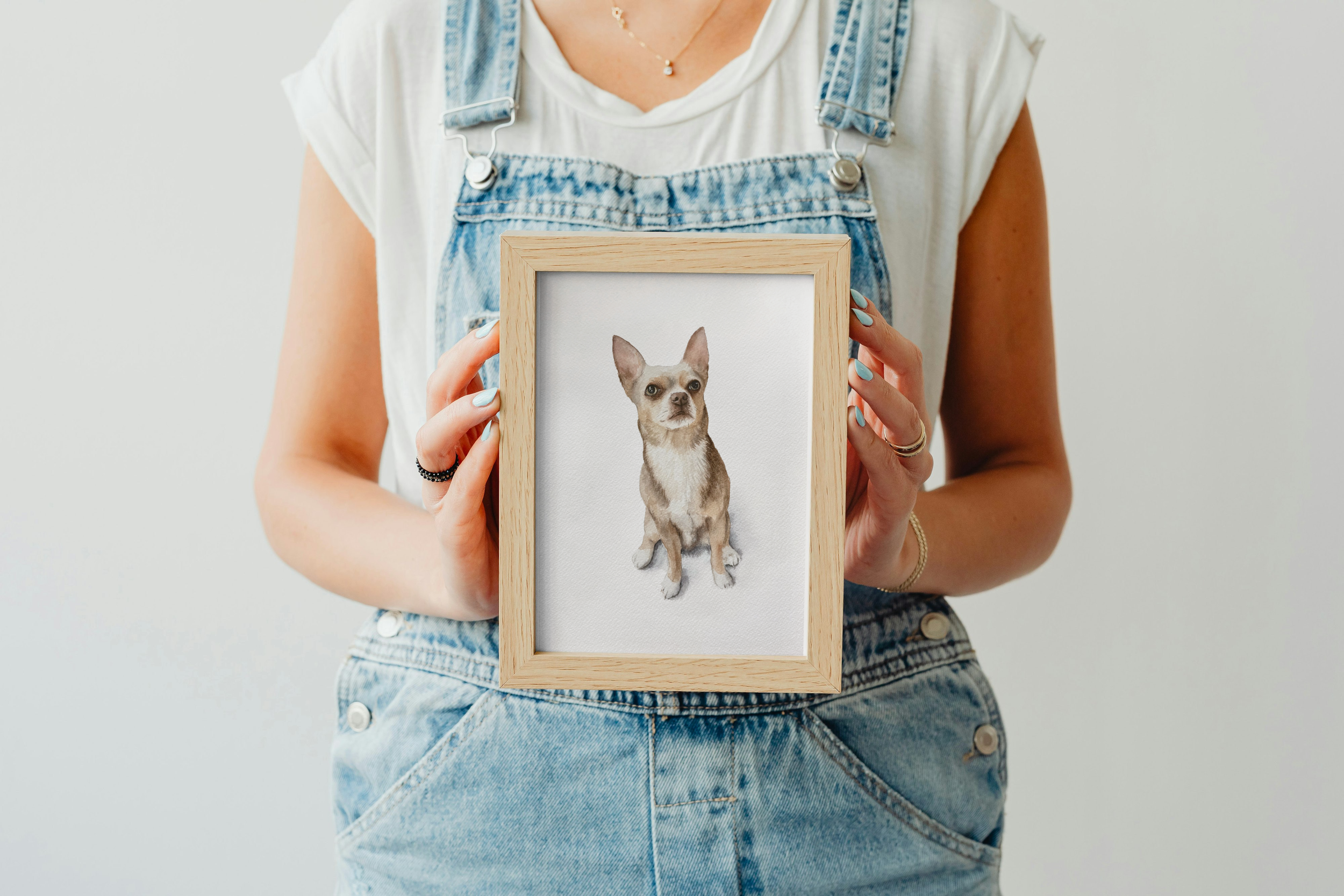 Person holding a framed watercolor portrait of a small dog, wearing a white t-shirt and denim overalls with a light background.
