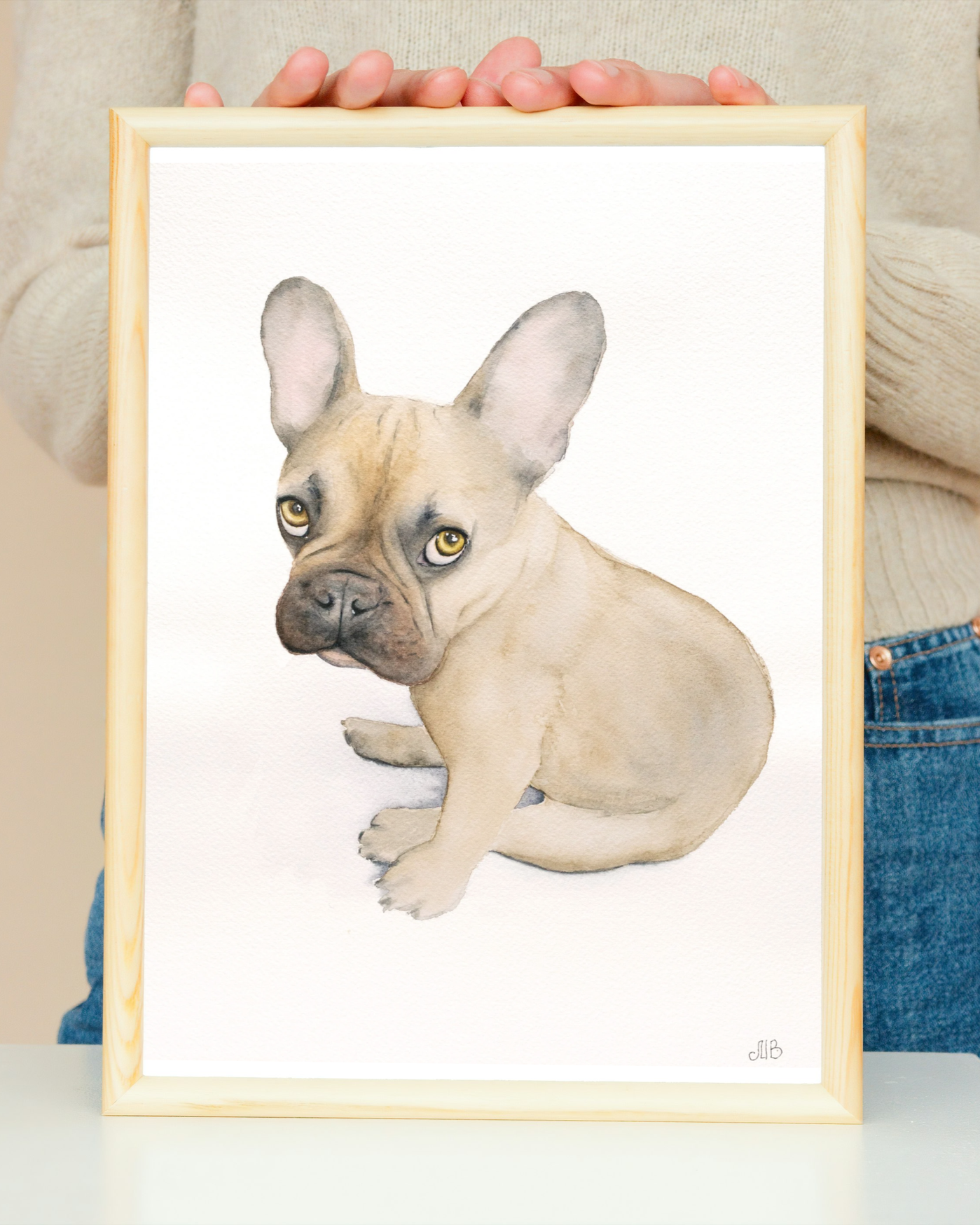 Watercolor painting of a sitting French Bulldog puppy with a tan coat and large ears, held in front of a beige background.