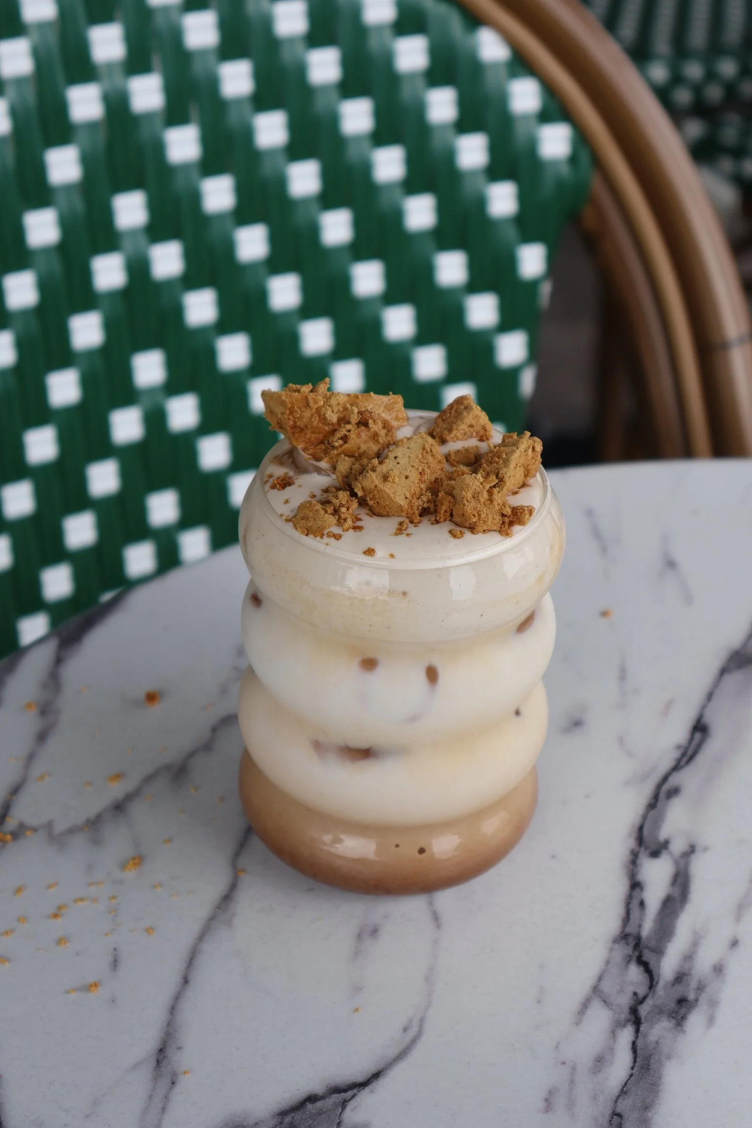 A layered dessert in a wavy glass, topped with crushed cookies, on a marble table with a green and white patterned chair in the background.