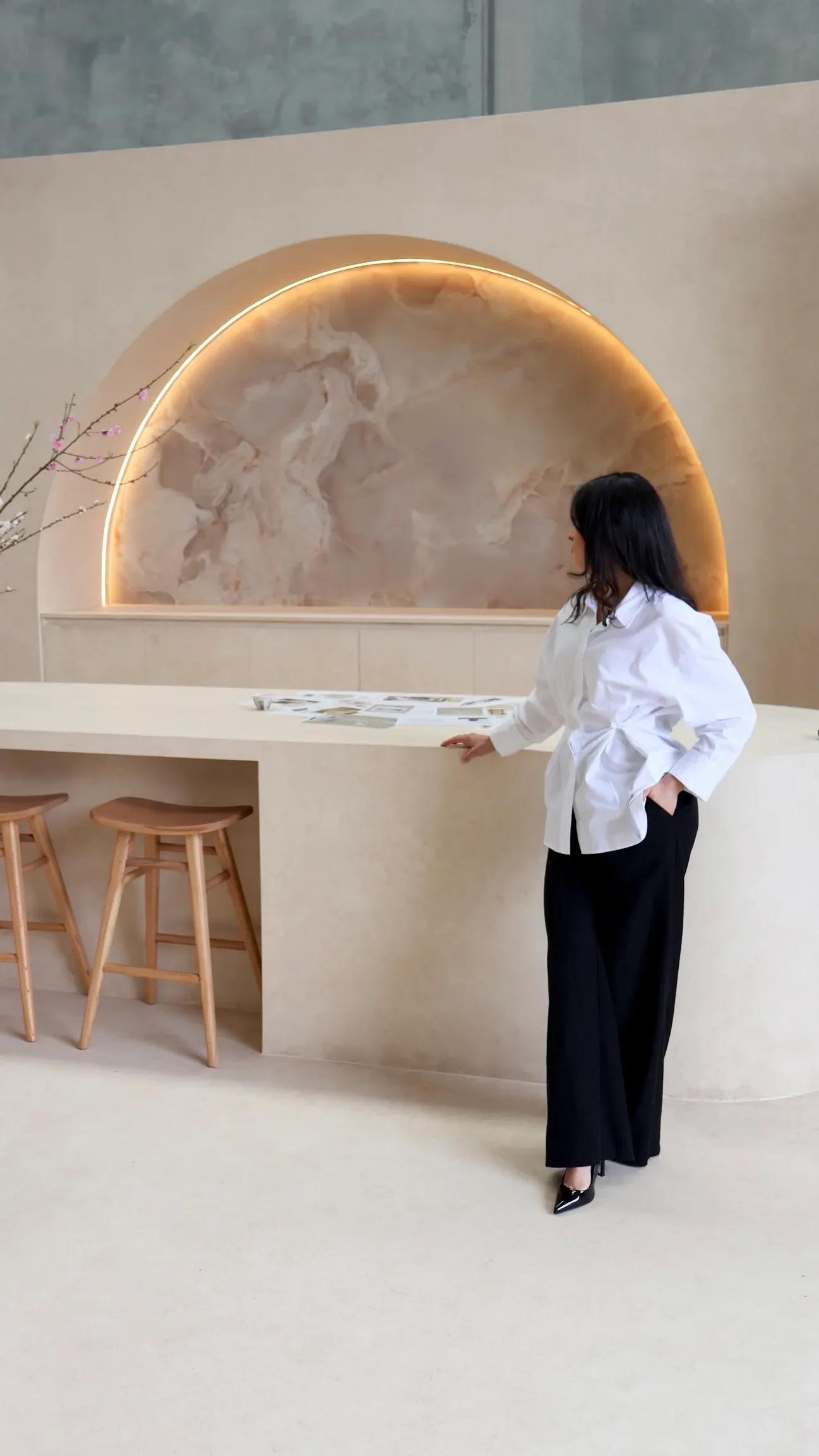 A woman in a white blouse and black wide-leg pants standing beside a beige marble countertop in a modern, minimalist interior with a decorative marble wall and built-in lighting in the background.