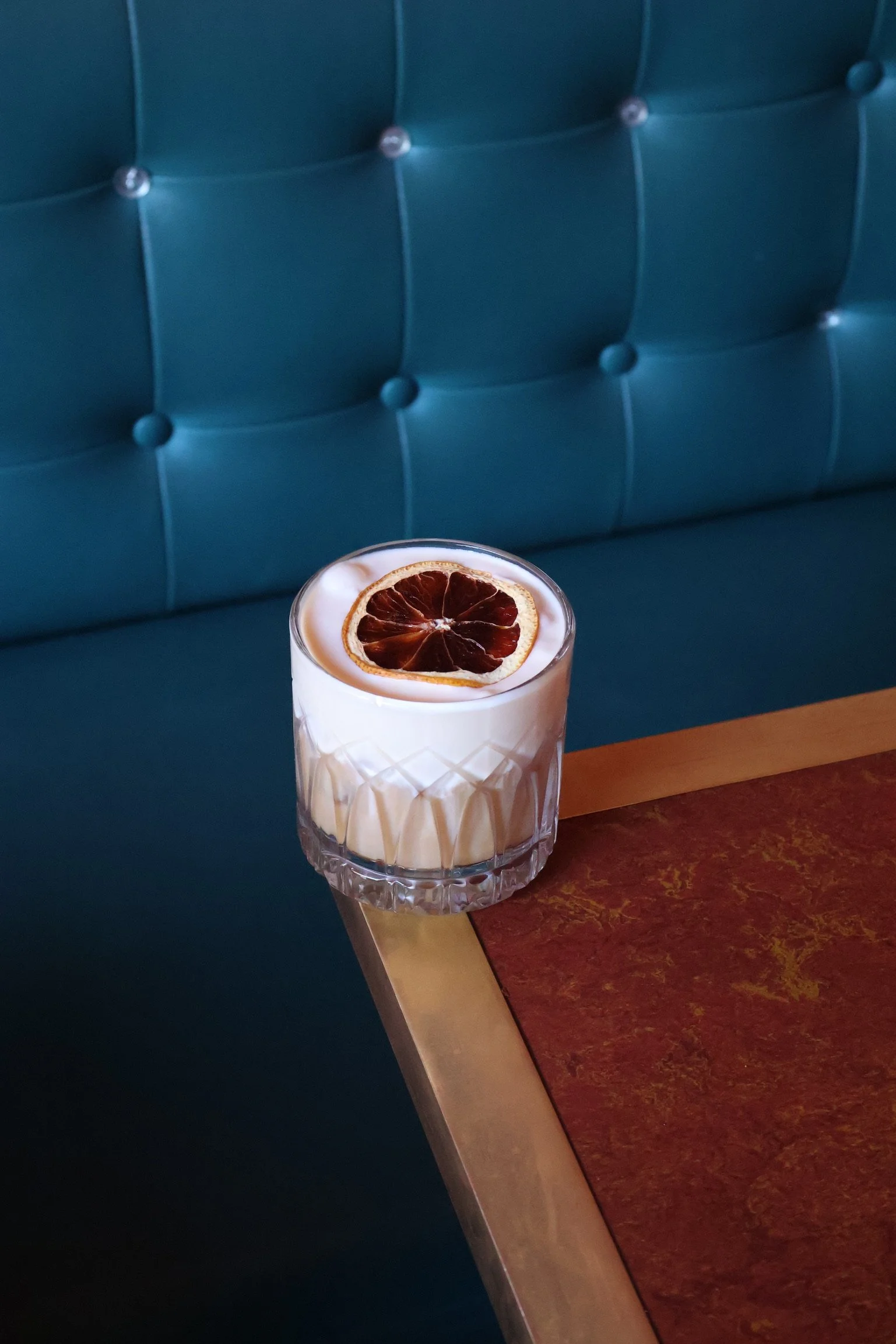 A glass of white cocktail garnished with a slice of dried blood orange, placed on a table with a blue cushioned booth in the background.