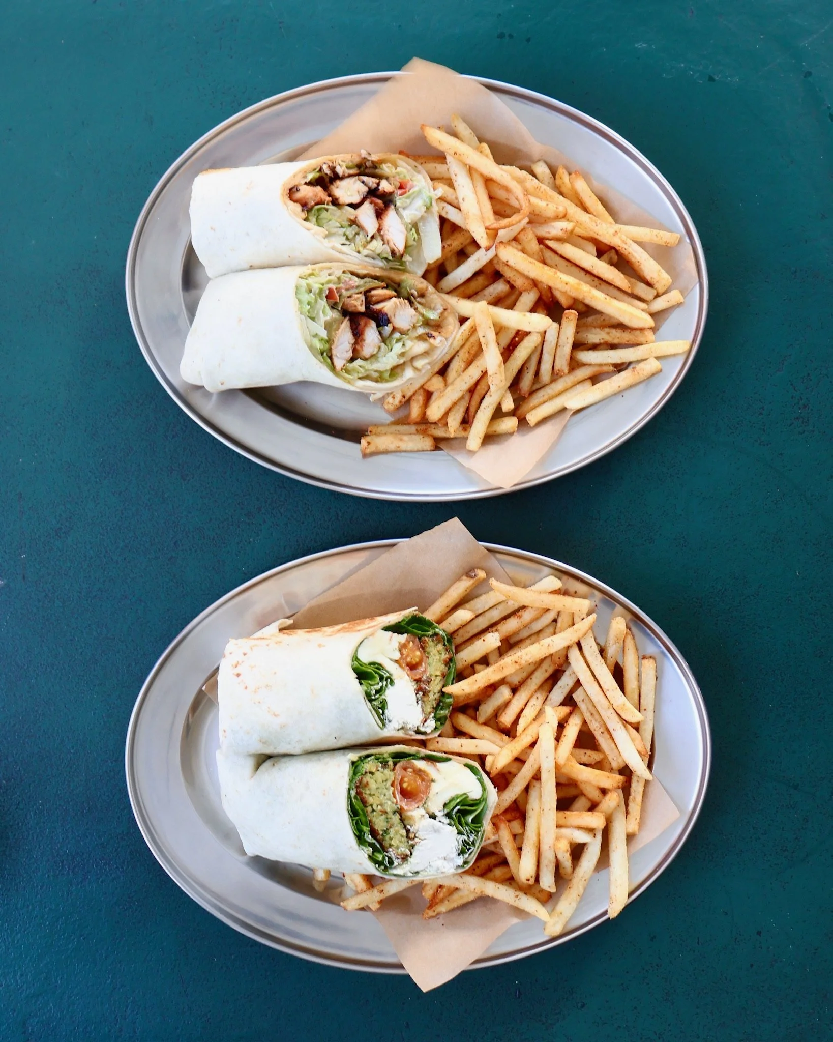 Two oval trays each containing a wrap sandwich and a portion of French fries on a green surface.