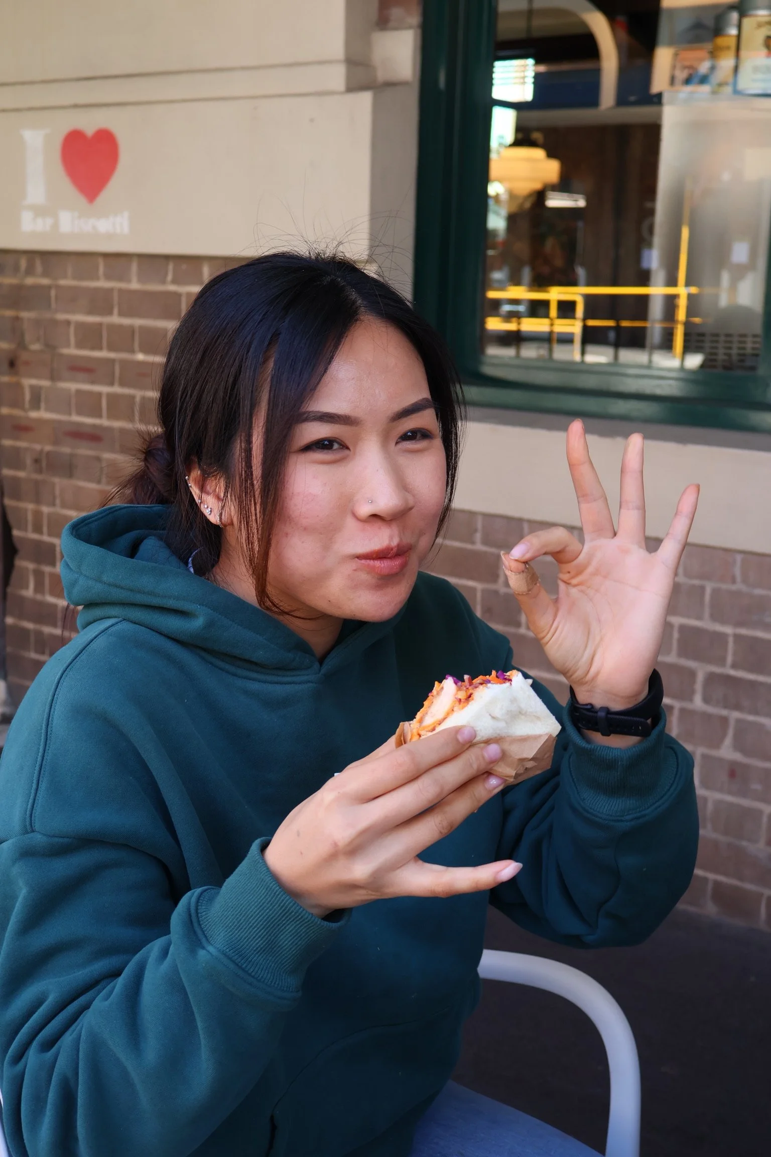 A young woman with dark hair and multiple earrings is sitting outside, wearing a teal hoodie, holding a sandwich with her right hand. She is making an 'okay' gesture with her left hand and looking at the camera with a slight smile. In the background, there is a brick wall, a window, and a sign that says 'I love Bar Biccetti' with a red heart.