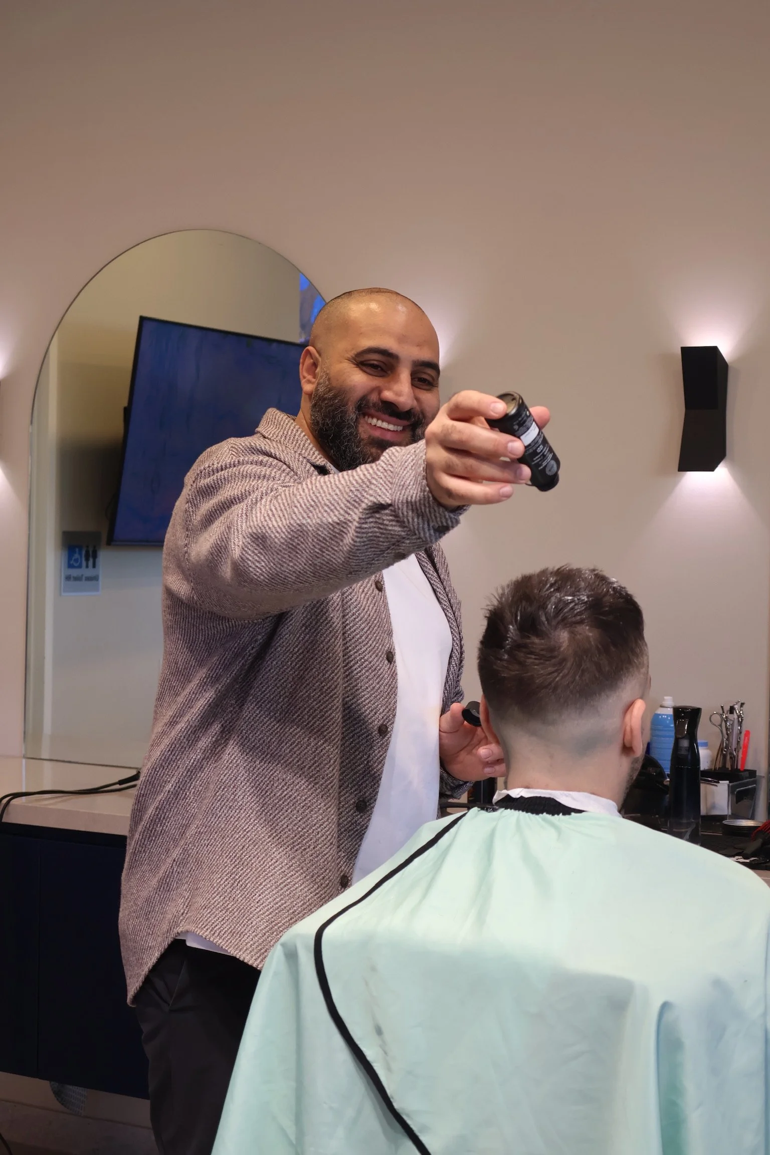 Barber with beard and bald head smiling, holding a spray bottle, cutting a young man's hair in a barber shop.