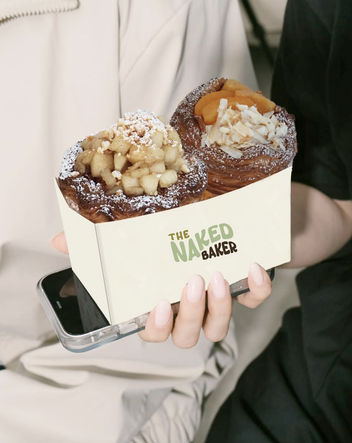 Two pastries with toppings in a box labeled 'The Naked Baker' being held by a person.
