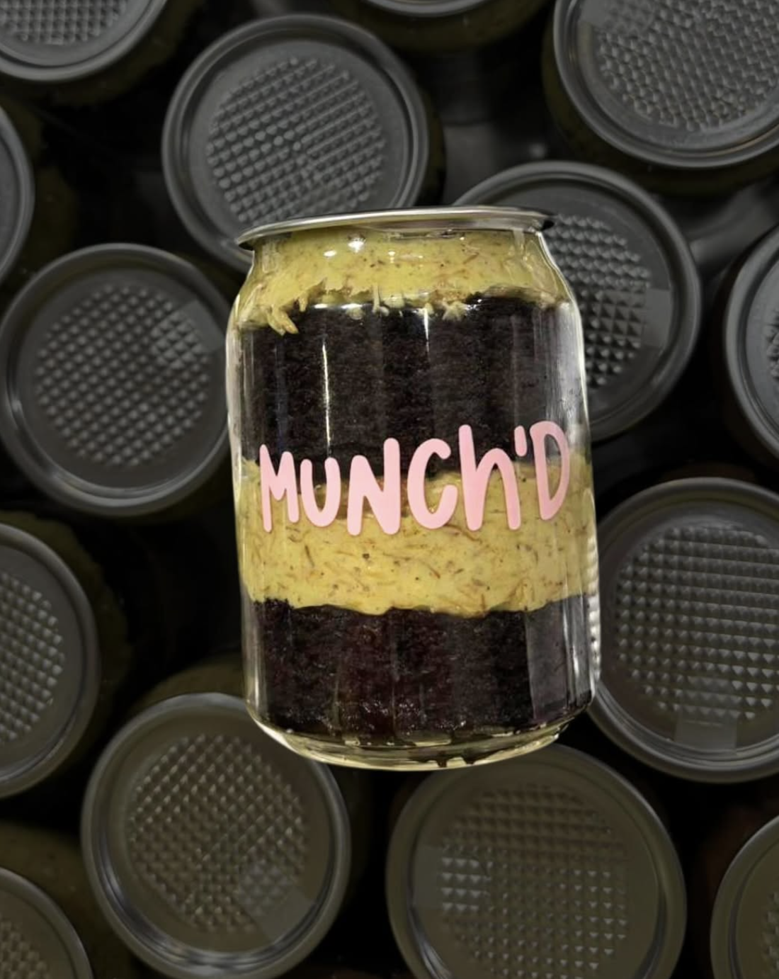 A glass jar with layers of yellow mustard and dark brown or black meat or antler slices, labeled 'MUNCH'D' with pink text, surrounded by cans with gray lids.