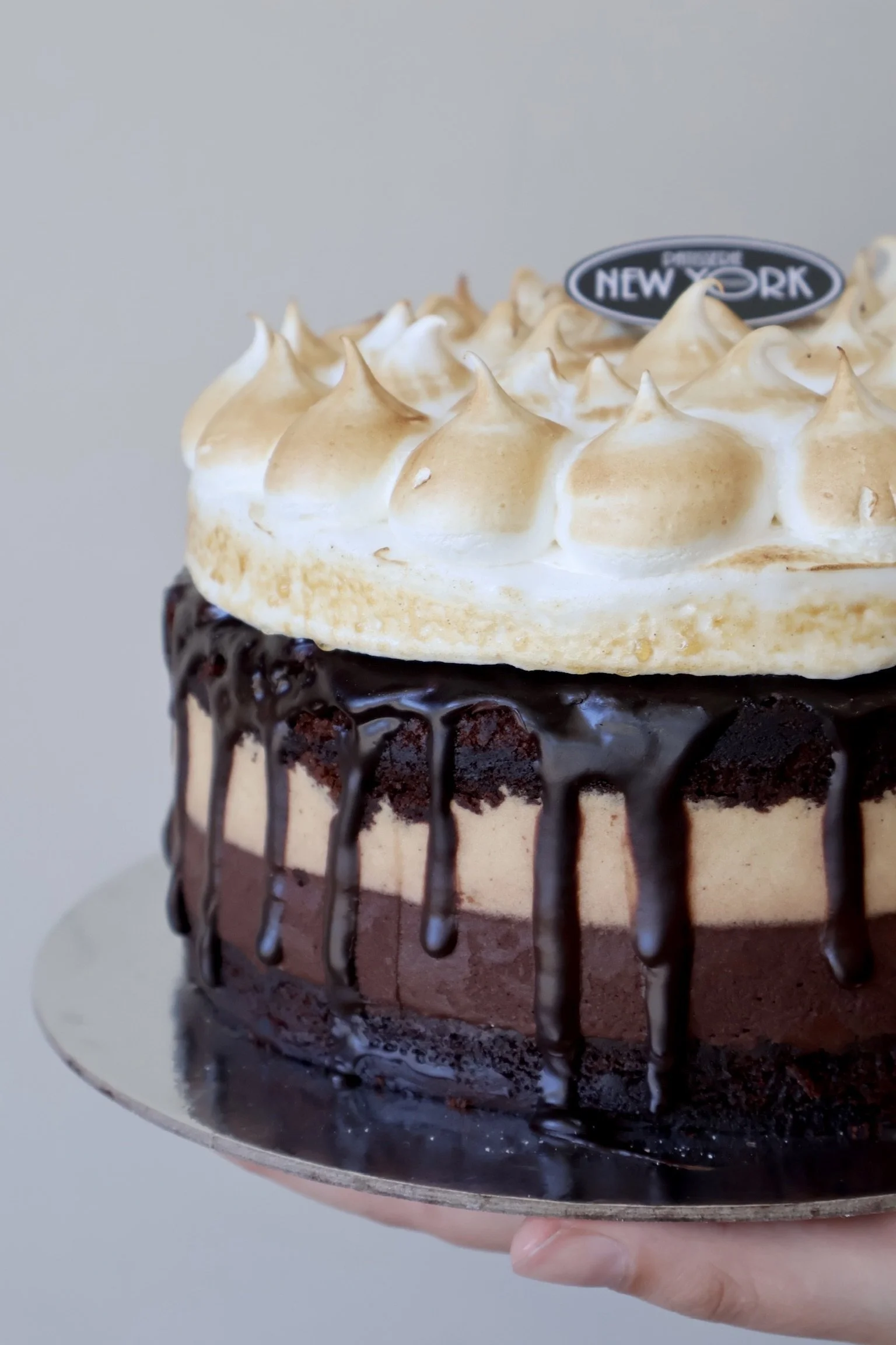 A close-up of a layered chocolate cake topped with toasted meringue peaks and a chocolate drizzle.